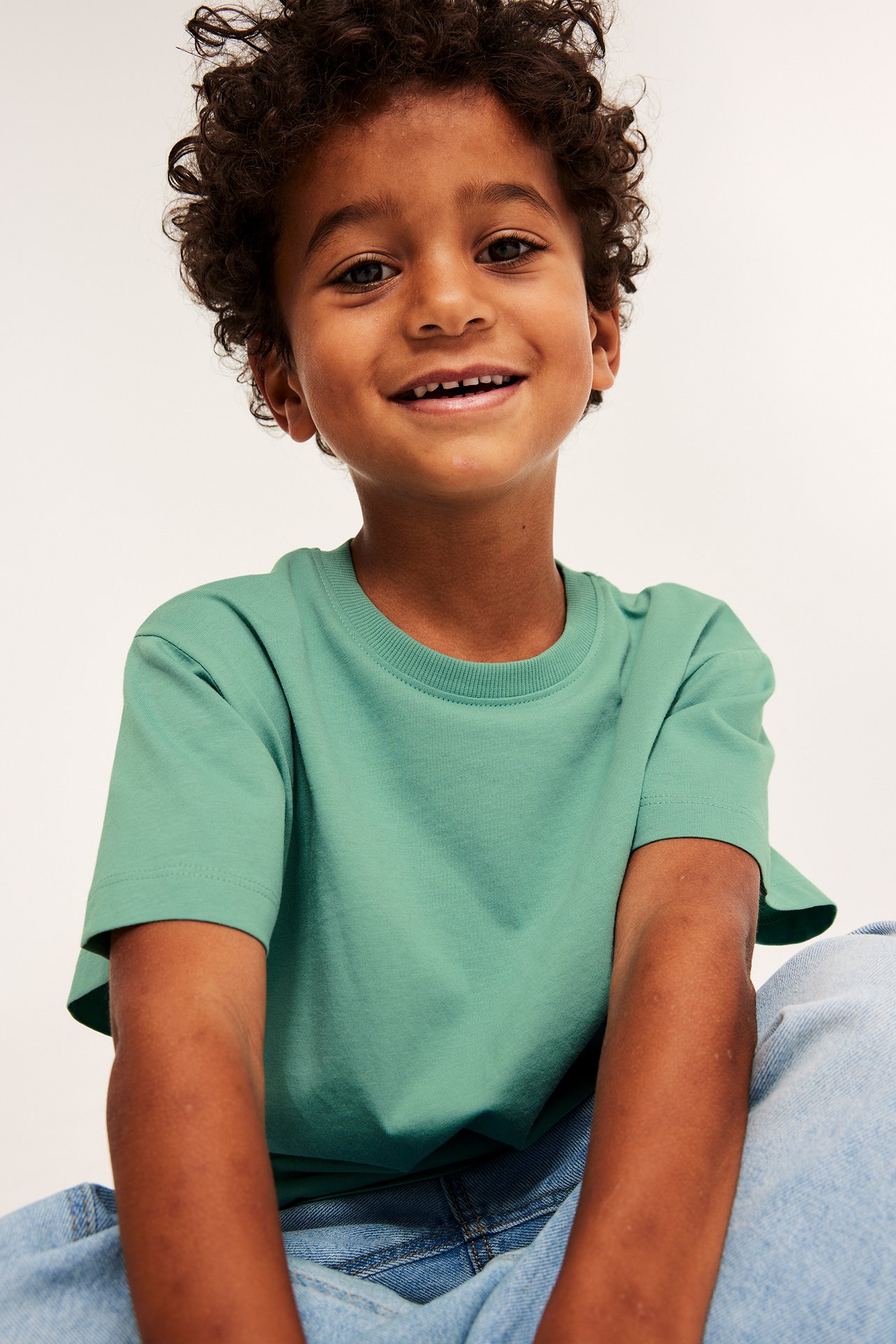 Frontalvisning av et smilende barn i en romslig, ensfarget grønn bomull T-skjorte og lyseblå jeans.