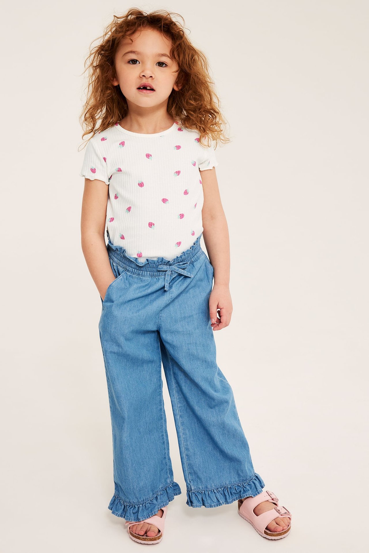 Framifrån: Ett barn i offwhite ribbstickad topp med jordgubbsmönster, blå vida byxor och rosa sandaler.