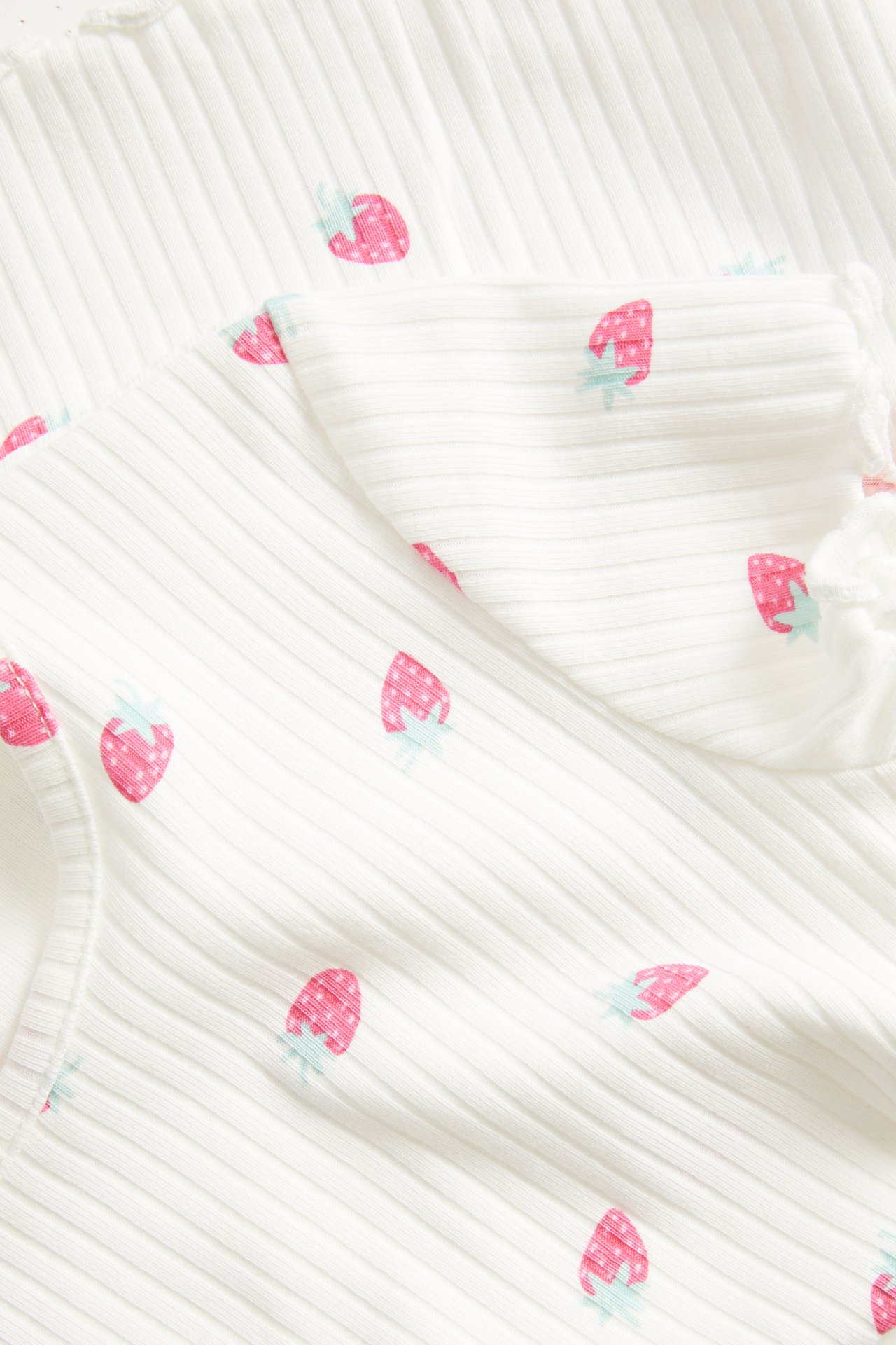 Lähikuva luonnonvalkoisesta ribbineulepaidasta, jossa vaaleanpunainen mansikkakuvio ja overlock-reuna.