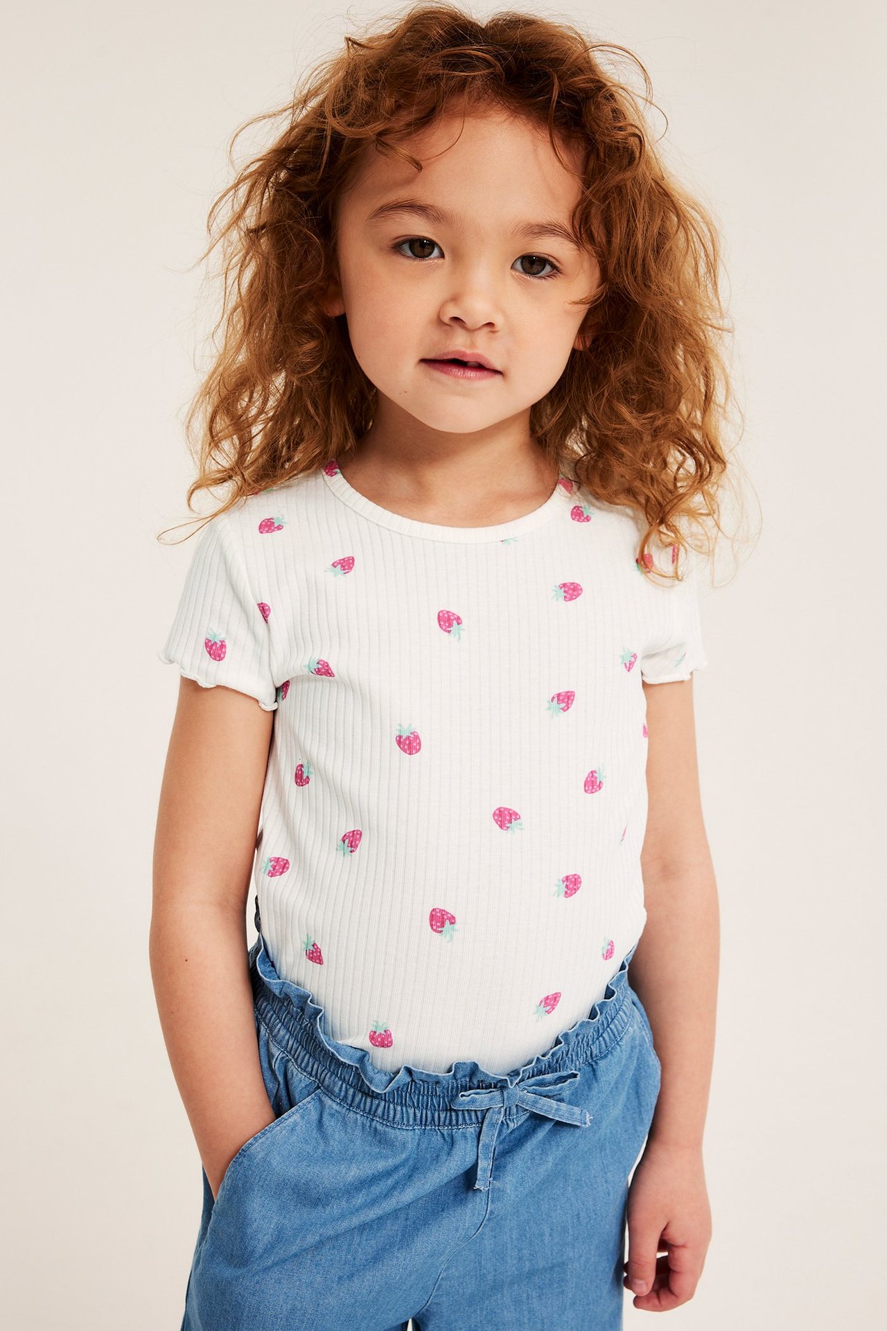 Edestä, lapsi luonnonvalkoisessa ribbipuuvillatopissa mansikkaprintillä ja sinisissä chambrayhousuissa.