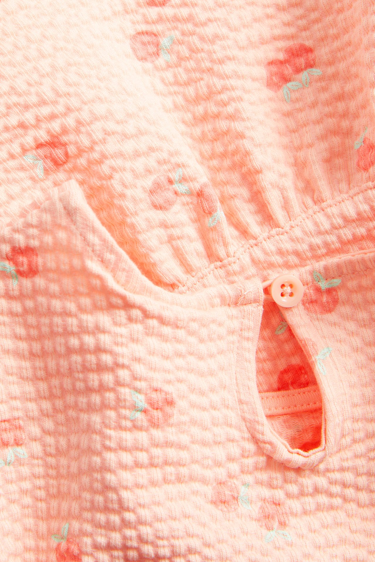 Zbliżenie na koralowy top peplum dla dziecka, z teksturowanego materiału z wzorem wiśni i guzikiem z tyłu.