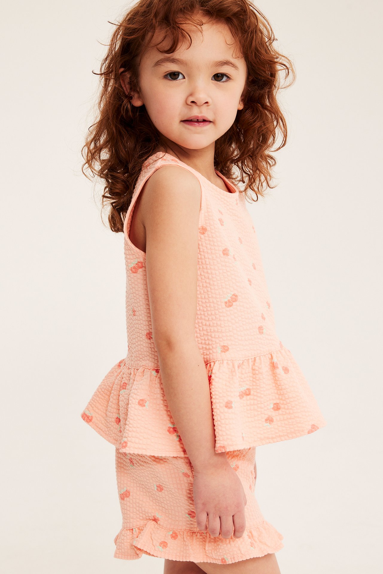 Widok z boku dziecka w koralowym topie peplum z wiśniami i pasujących szortach z falbanką.