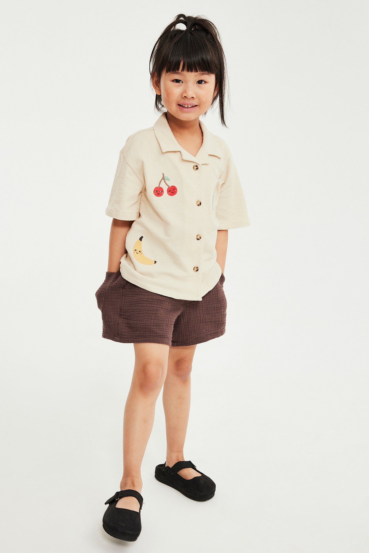 Edestä: Lapsi yllään beige lyhythihainen puuvillapaita hedelmäkirjonnalla, ruskeat kuvioidut shortsit ja mustat kengät.