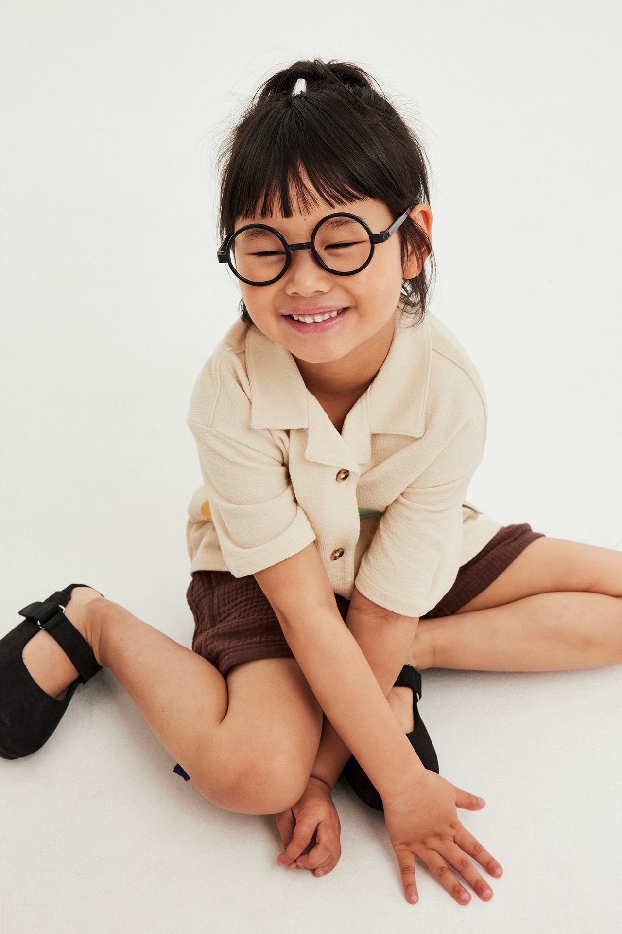 Framsida, leende barn i beige kortärmad trikåskjorta, bruna shorts, svarta skor och runda glasögon.