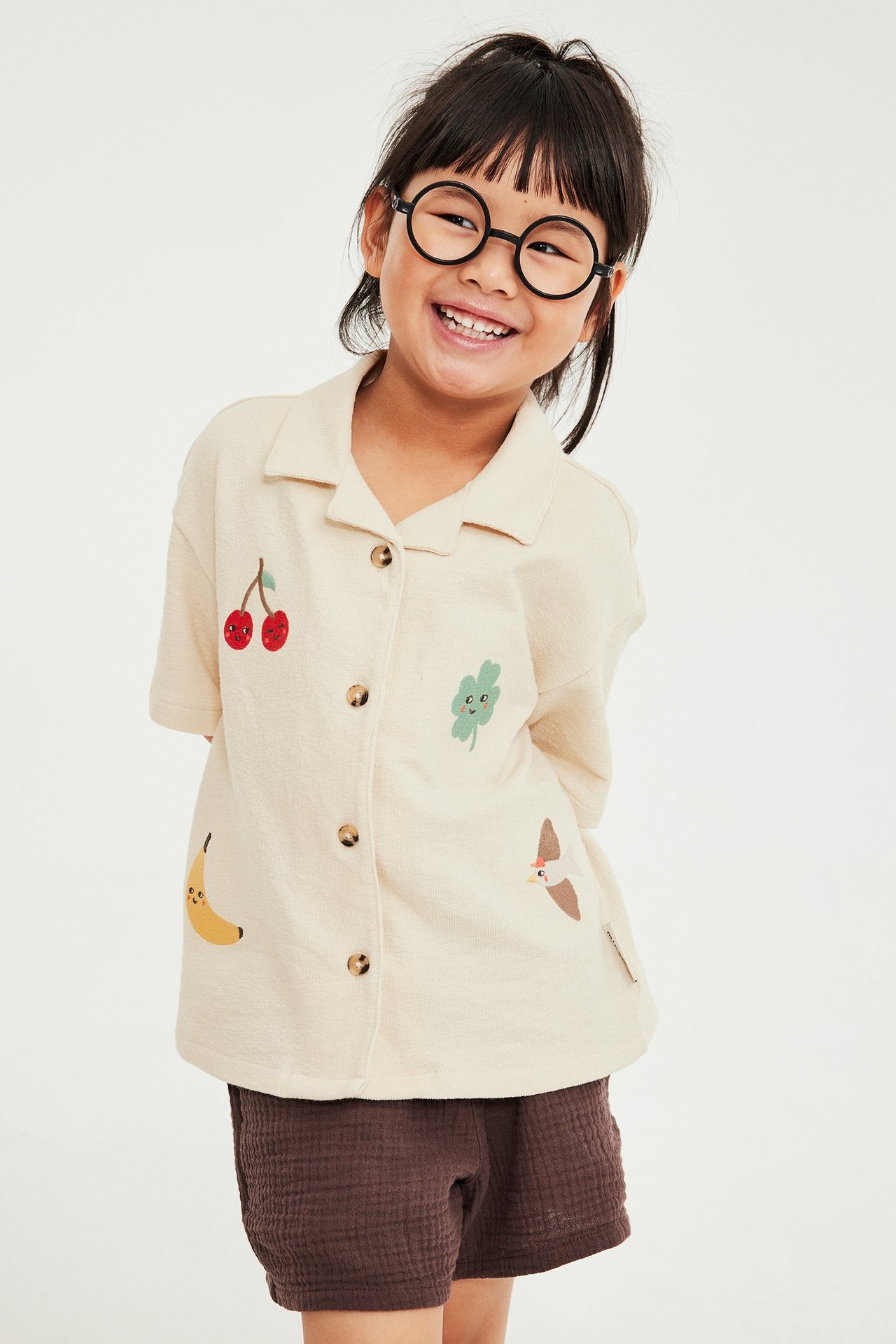 Framifrån: Barn i beige broderad bomullsskjorta, bruna shorts och svarta runda glasögon.