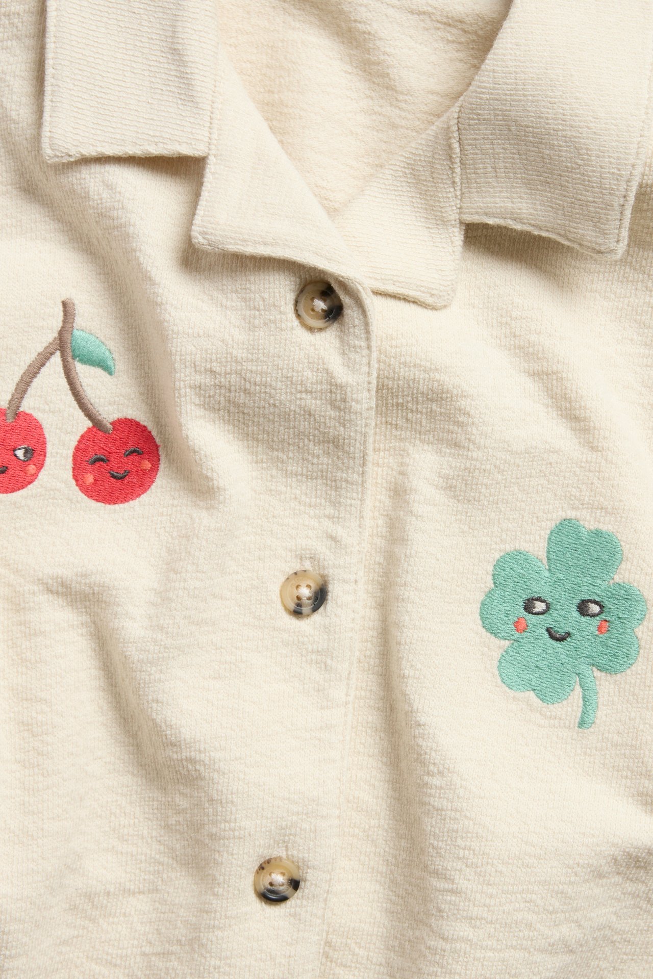 Framifrån: Barnskjorta i beige bomullstrikå med knappar, broderade glada körsbär och en grön klöver.