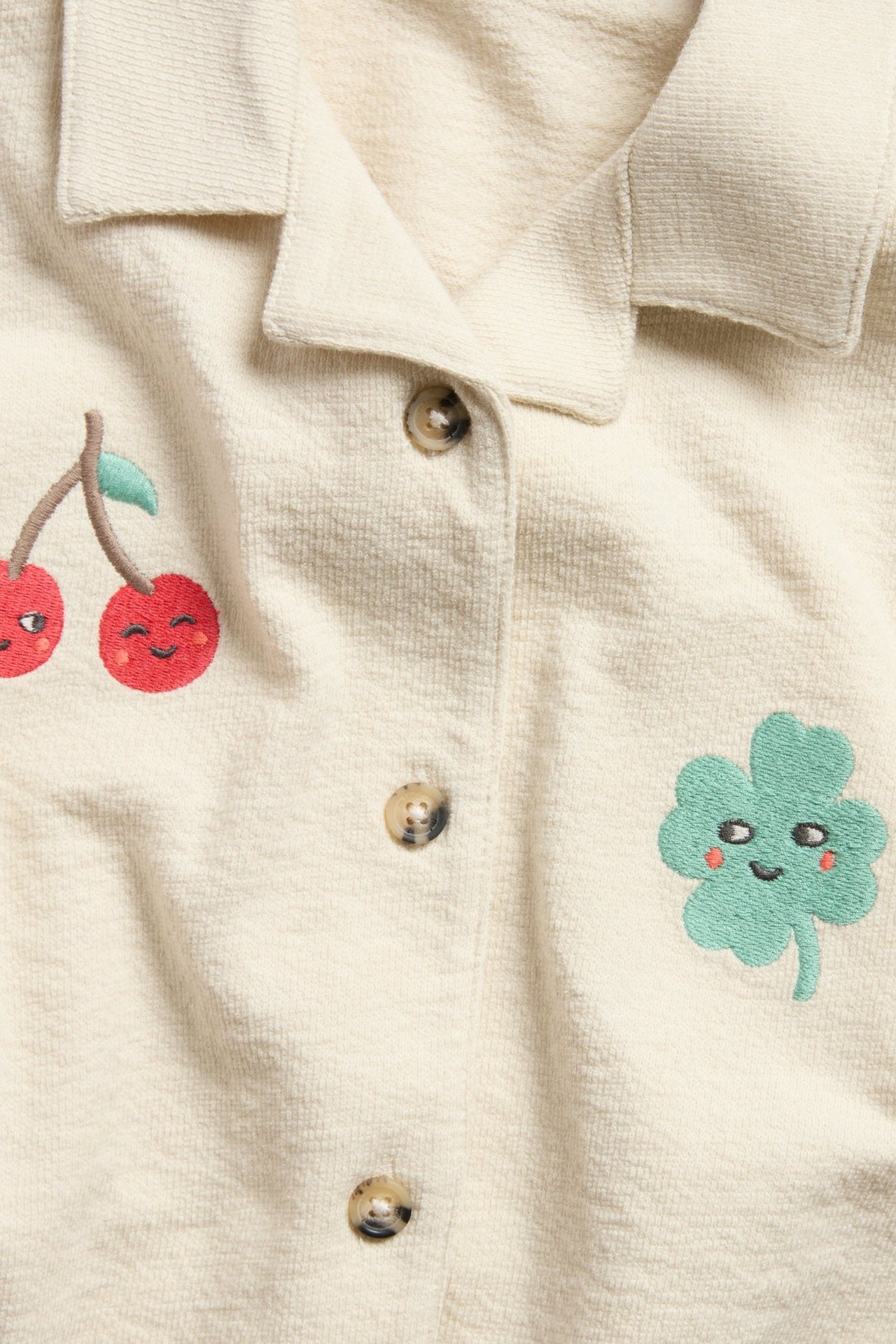 Widok z przodu: Beżowa koszula dziecięca z dzianiny bawełnianej z guzikami, haftowanymi wisienkami i zieloną koniczyną.