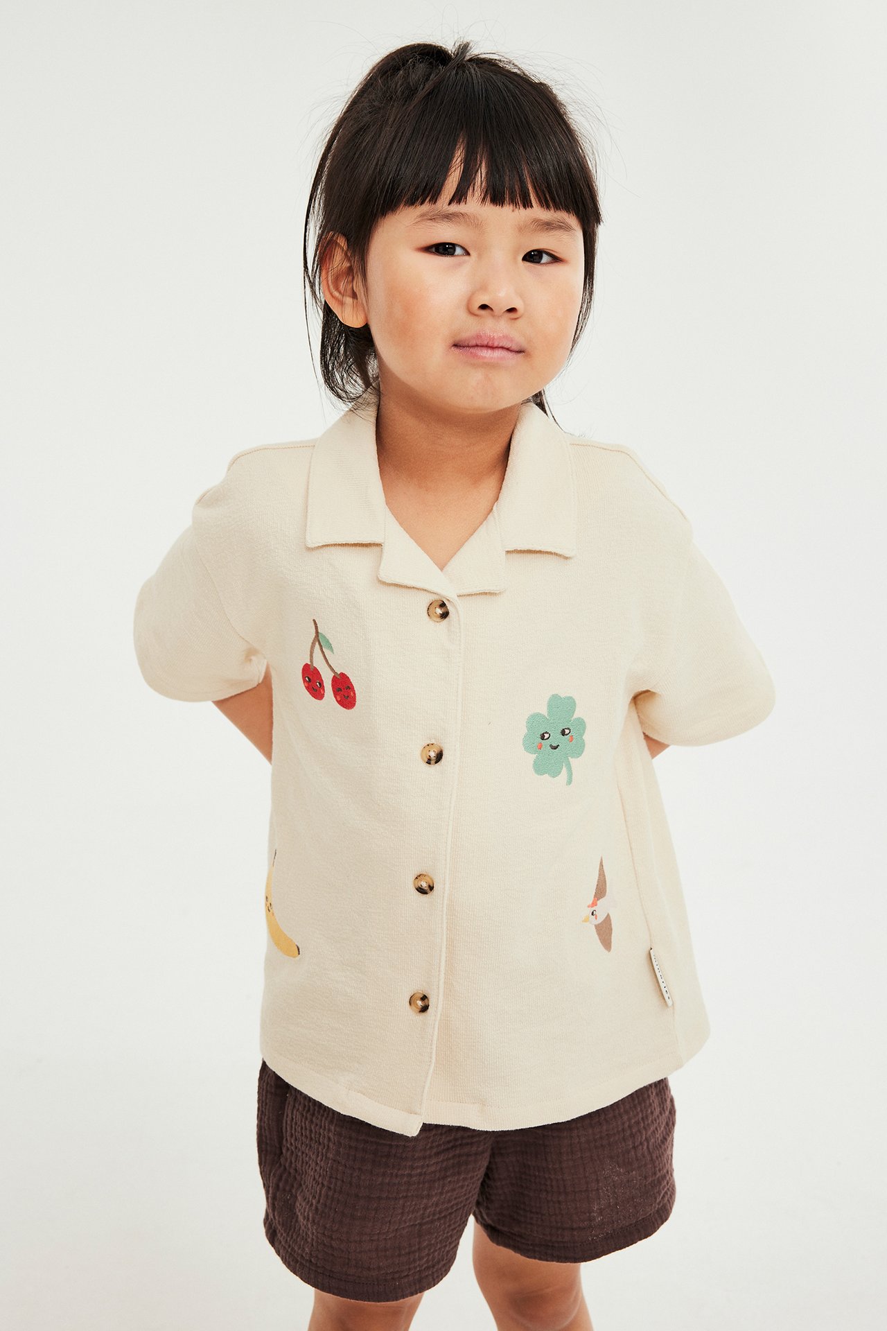 Widok z przodu dziecka w beżowej bawełnianej koszuli z krótkim rękawem z kolorowymi aplikacjami i brązowych szortach.