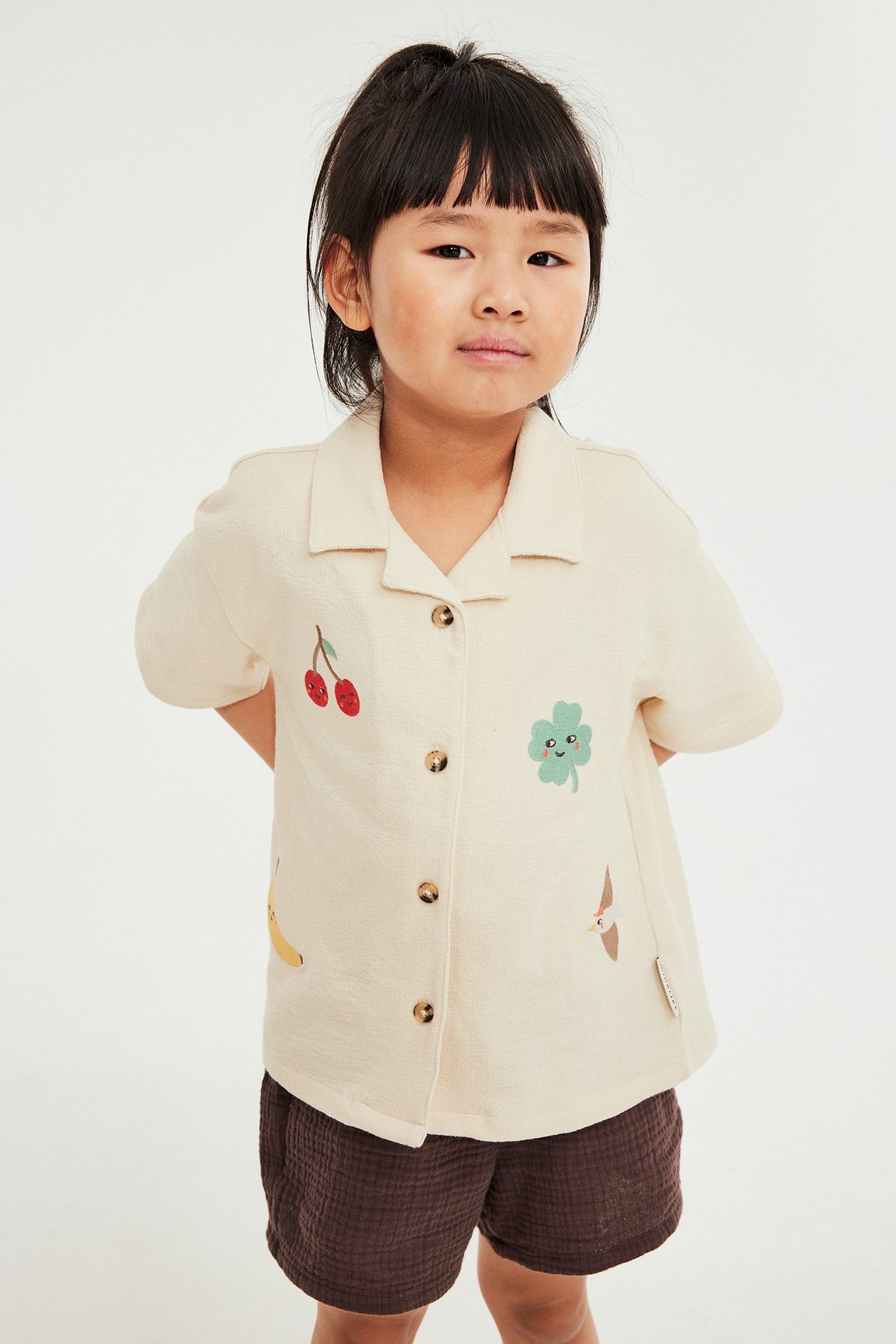 Widok z przodu dziecka w beżowej bawełnianej koszuli z krótkim rękawem z kolorowymi aplikacjami i brązowych szortach.
