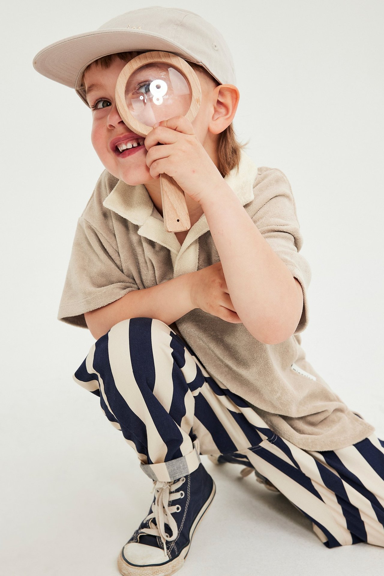 Frontvy: Ett barn bär en beige kortärmad frottétröja med krage, mörkblårandiga byxor, beige keps och blå sneakers.