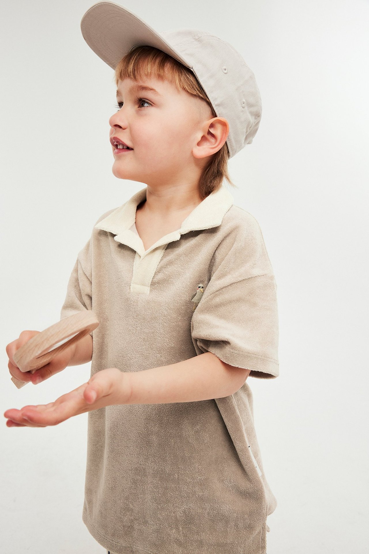 Sidovy av ett barn i en beige frottépiquétröja med benvit krage, djurbrodyr och en ljusbeige keps.