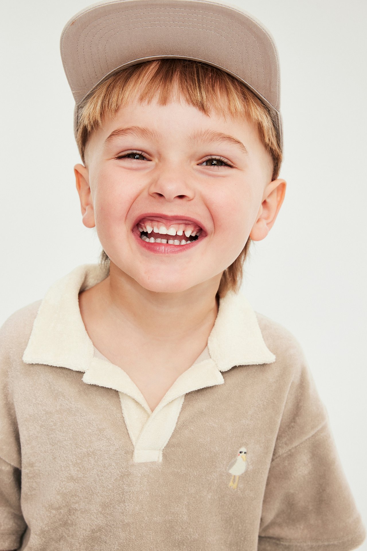Frontvy av ett skrattande barn i beige kortärmad frottétröja med krämkrage, fågelbrodyr och beige keps.