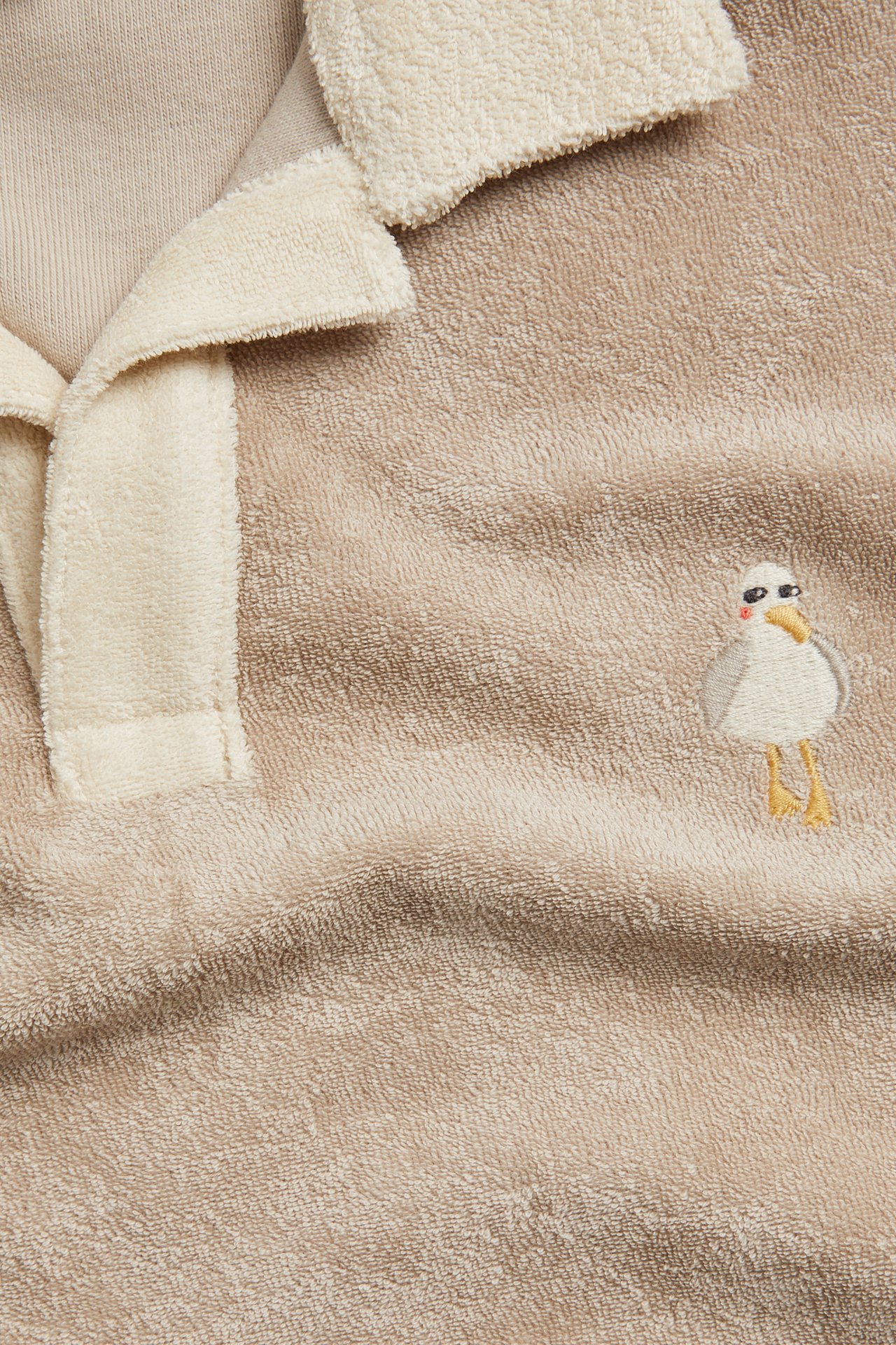 Närbild framifrån på en beige kortärmad frottétopp med gräddvit krage och broderad mås.