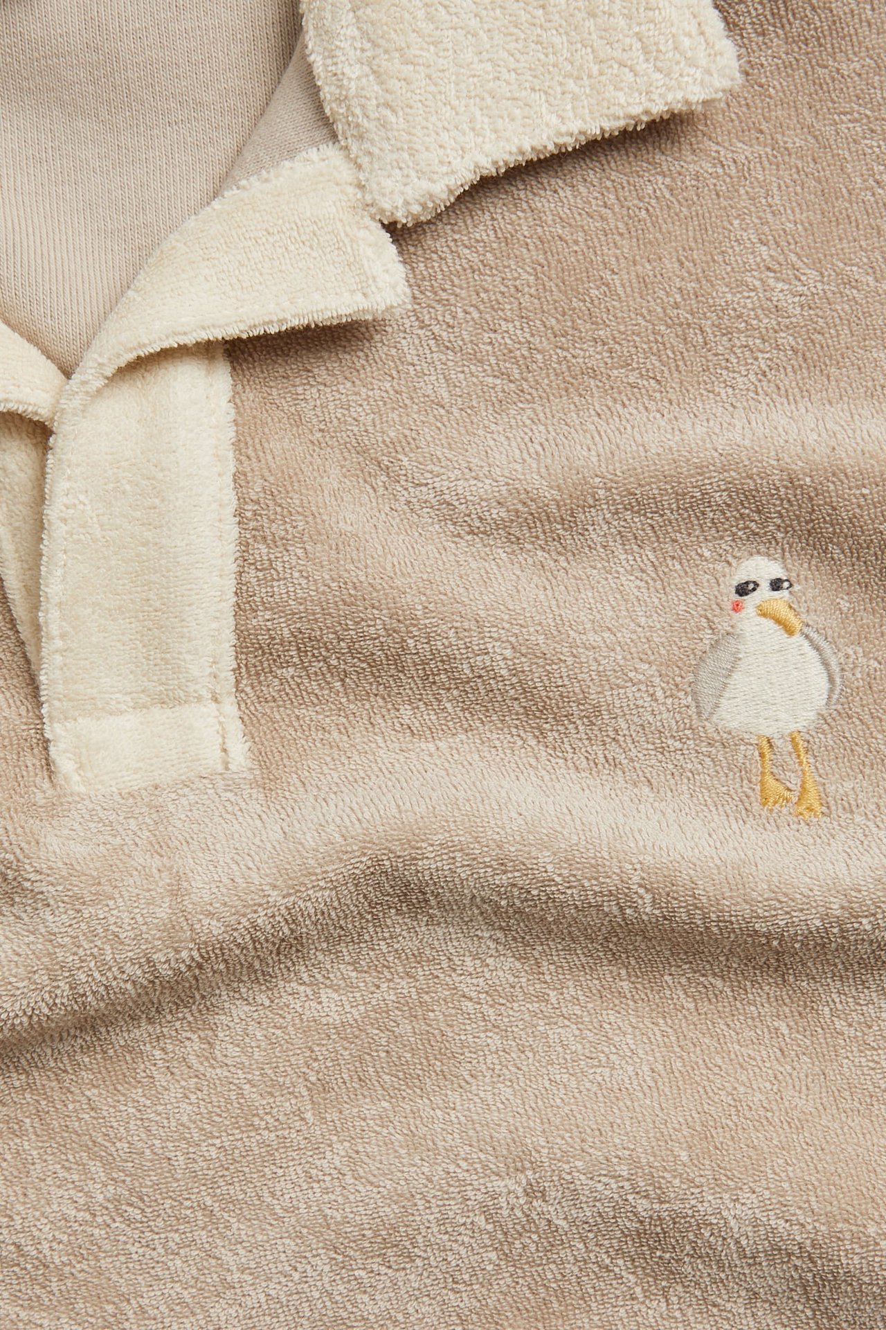 Zbliżenie przód beżowego, krótkiego topu frotte z kremowym kołnierzykiem i haftowaną mewą.