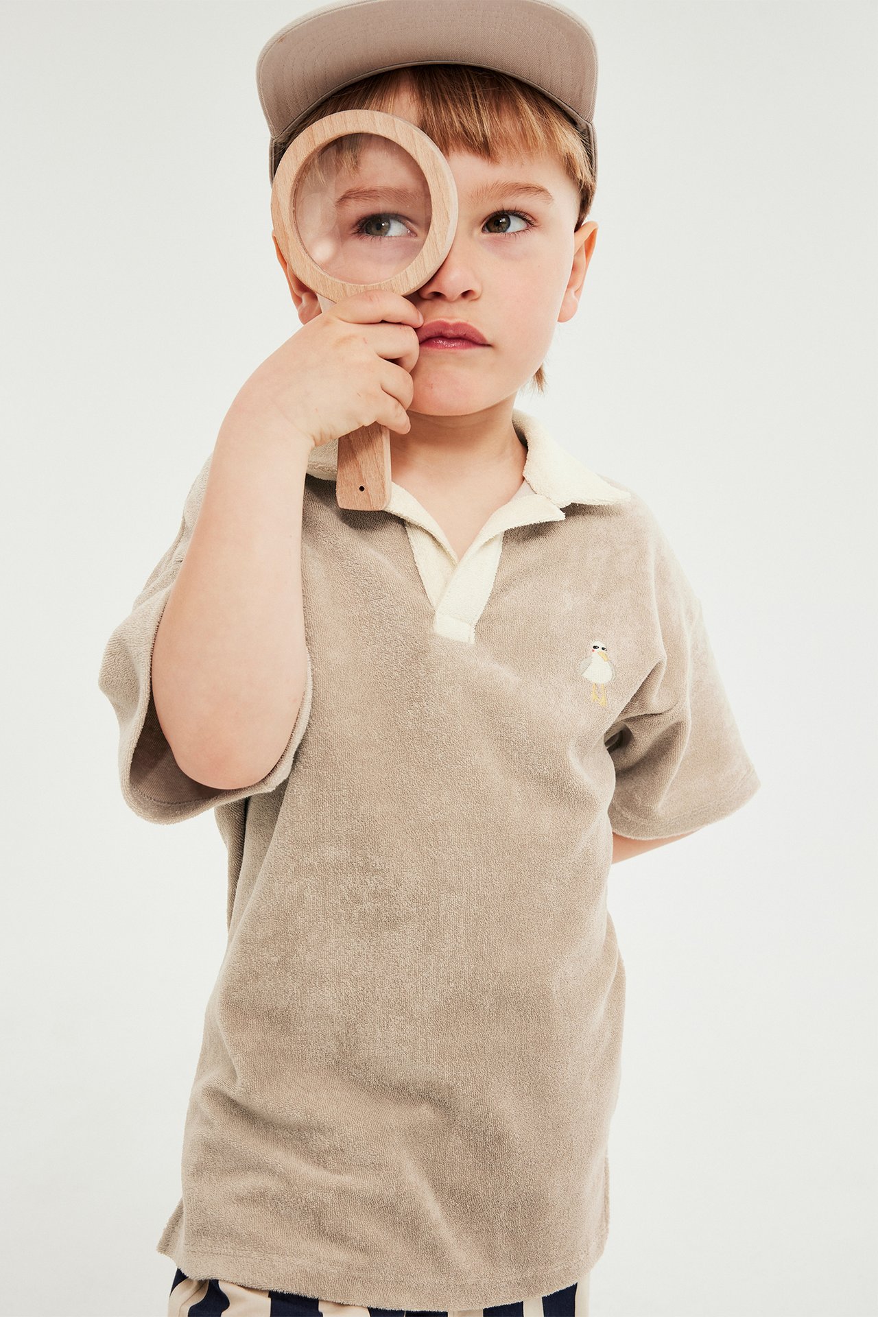 Barn i beige kortärmad frottépolo med fågelbrodyr, ljus krage, beige keps, håller förstoringsglas.