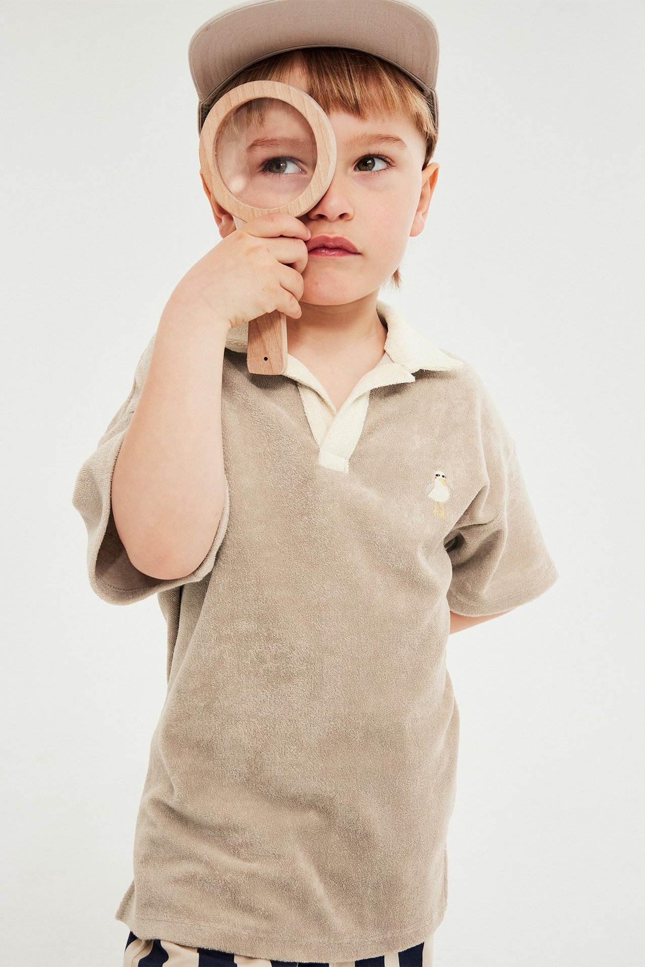 Dziecko w beżowym frotowym polo z haftem ptaka, jasnym kołnierzykiem, beżowej czapce, trzyma lupę.