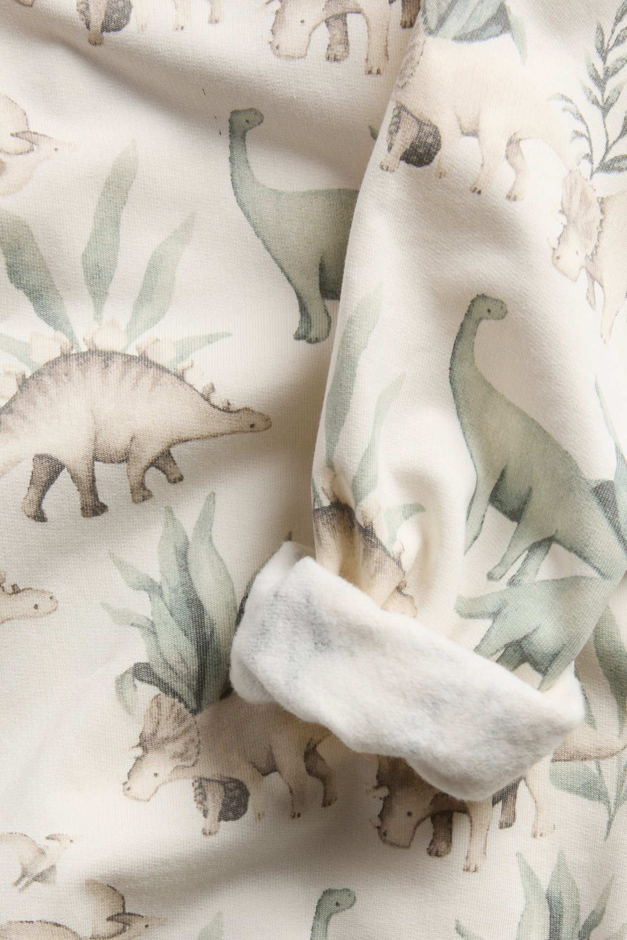 Lähikuva kermanvärisestä kankaasta, jossa on vihreä ja beige dinosaurus- ja lehtikuvio.