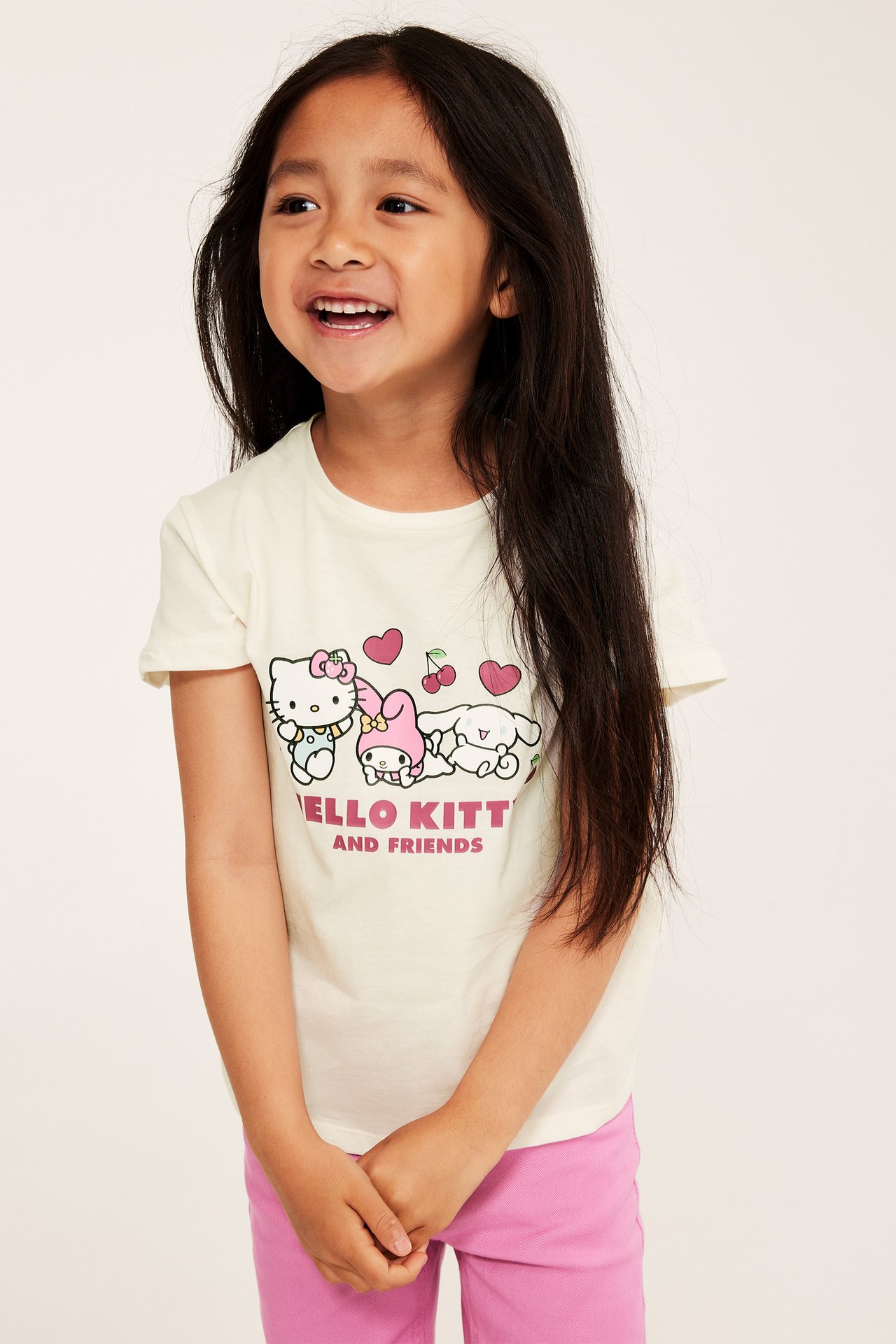 Framifrån: Ett barn i krämfärgad Hello Kitty-t-shirt med tryck och rosa byxor.