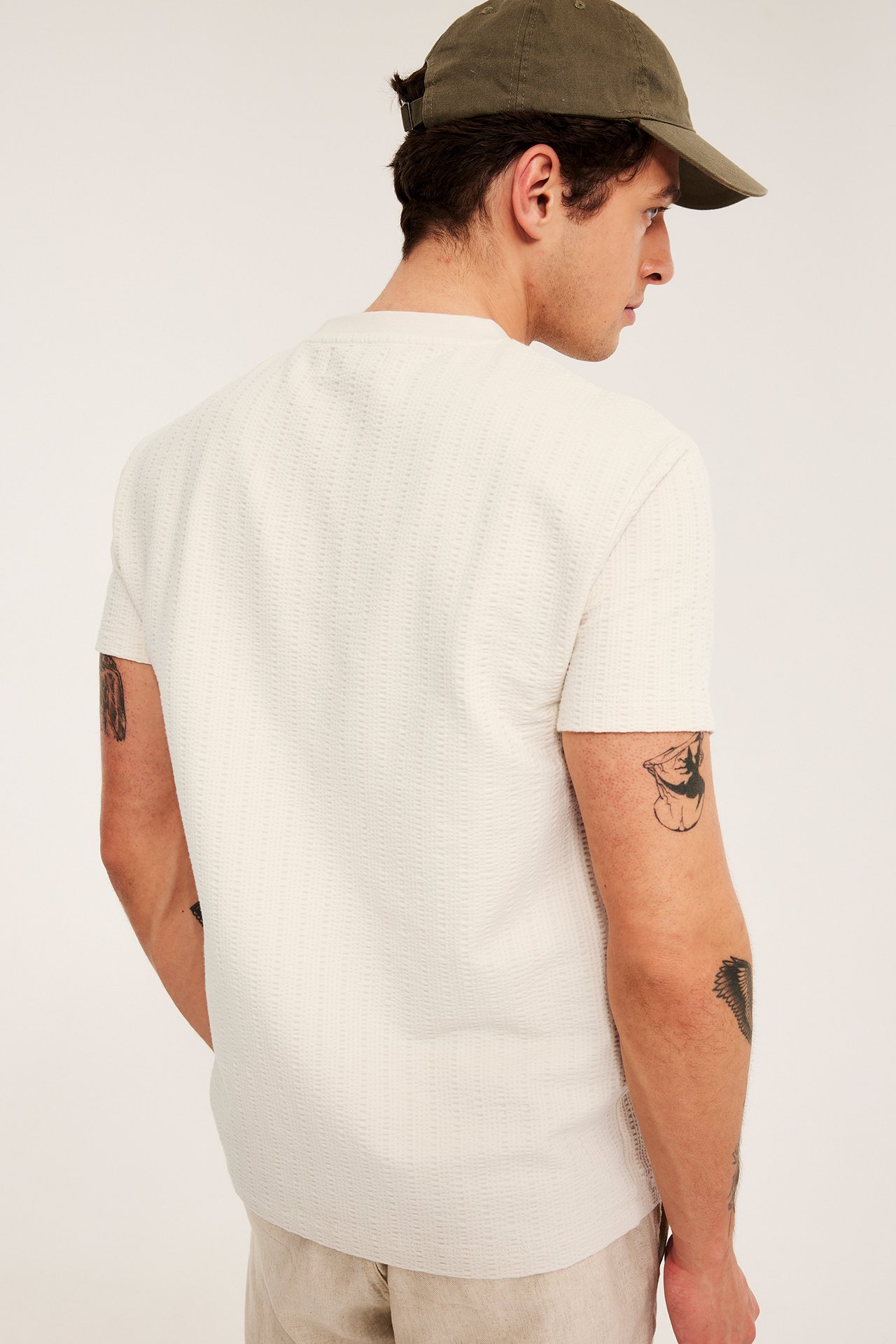 Man i ljusbeige strukturstickad t-shirt, olivgrön keps och ljusbeige shorts, sedd bakifrån.