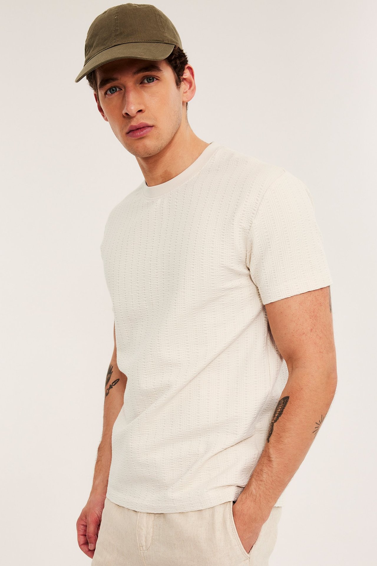 Edestäpäin: Mies yllään vaaleanbeige kuvioitu T-paita, vaaleanbeiget housut ja oliivinvihreä lippalakki.