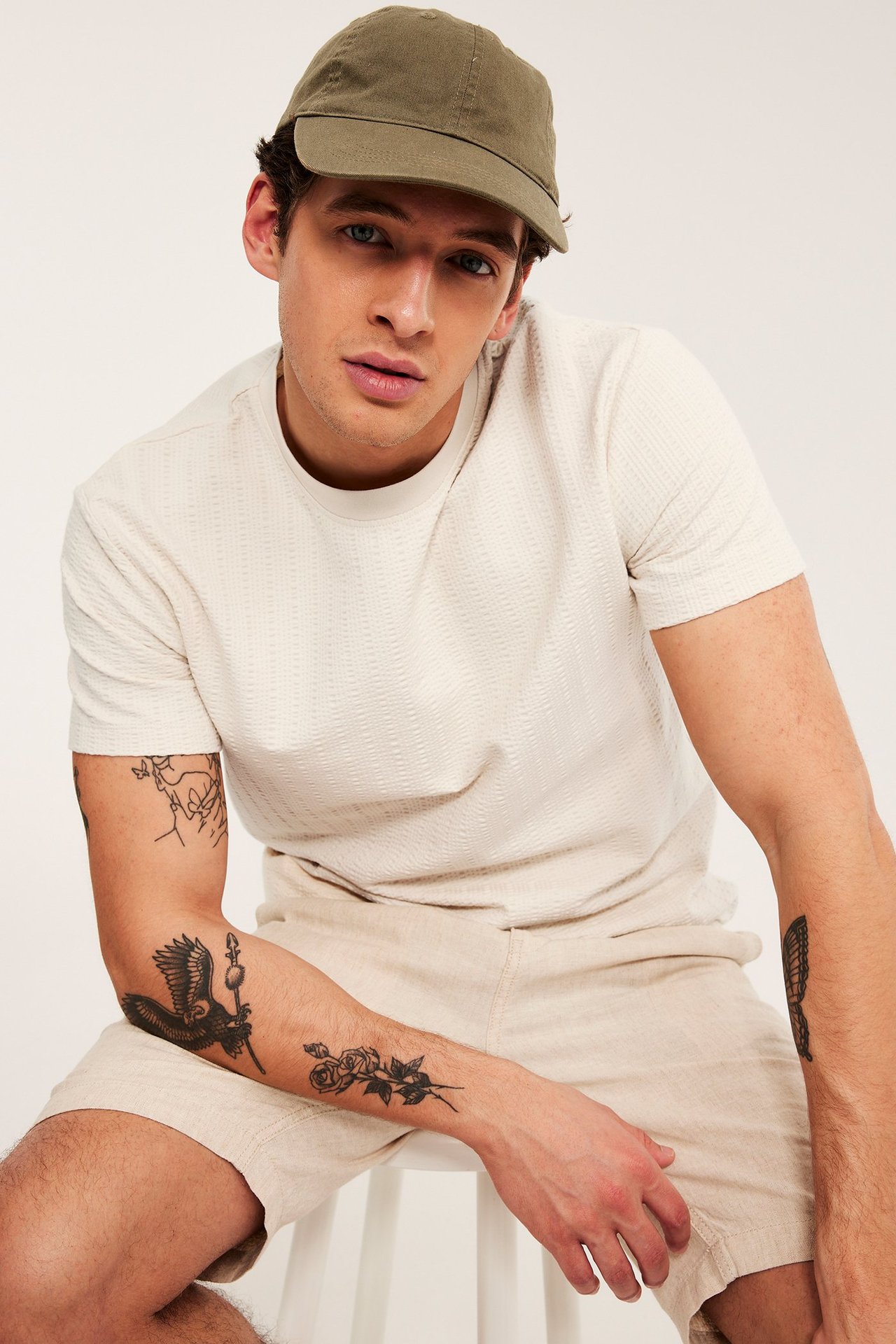 Frontvisning: Mann i lys beige strukturert t-skjorte, lyse beige shorts og en oliven grønn caps.