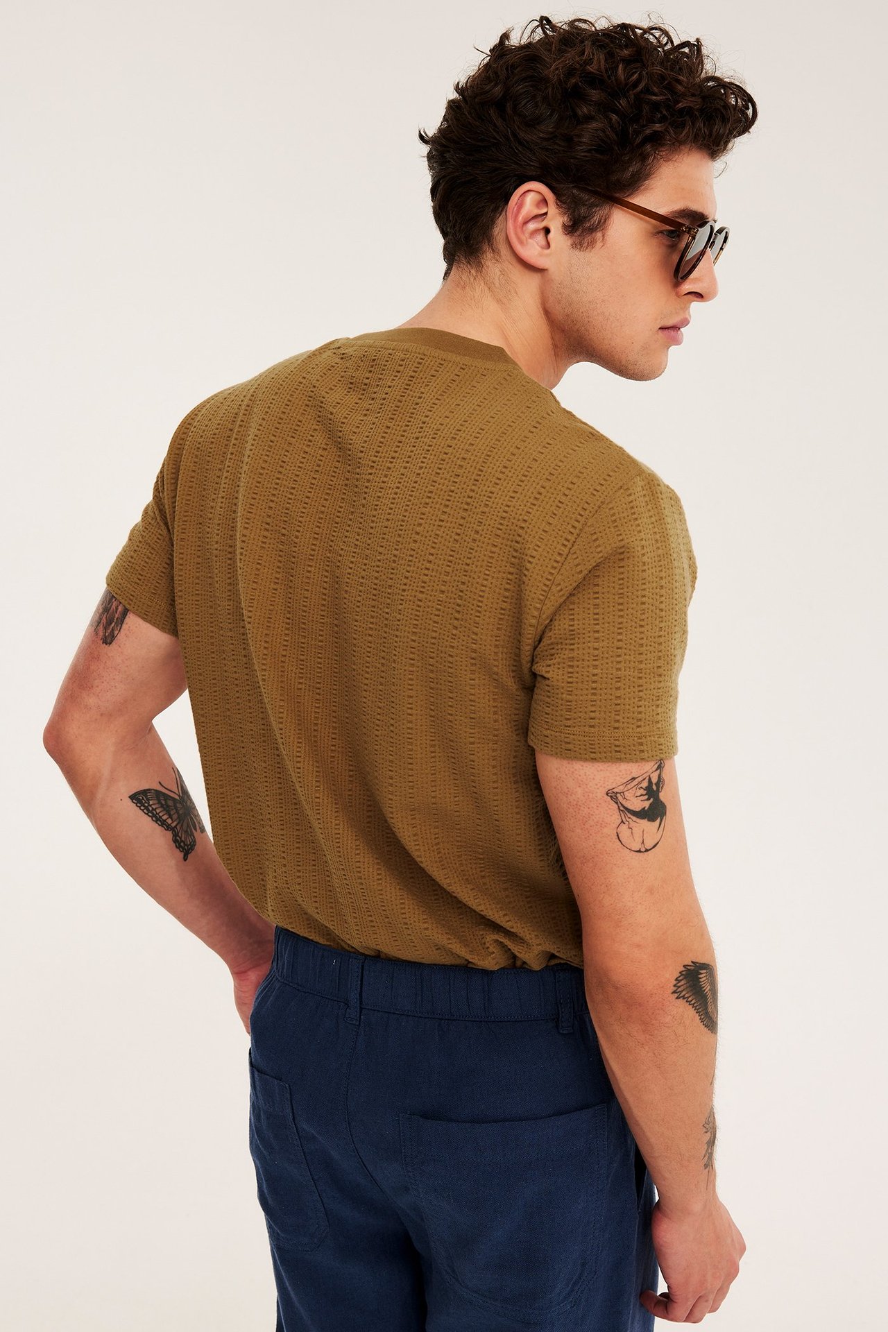 Man i brun O-ringad strukturstickad t-shirt, blå byxor och bruna solglasögon, bakifrån.