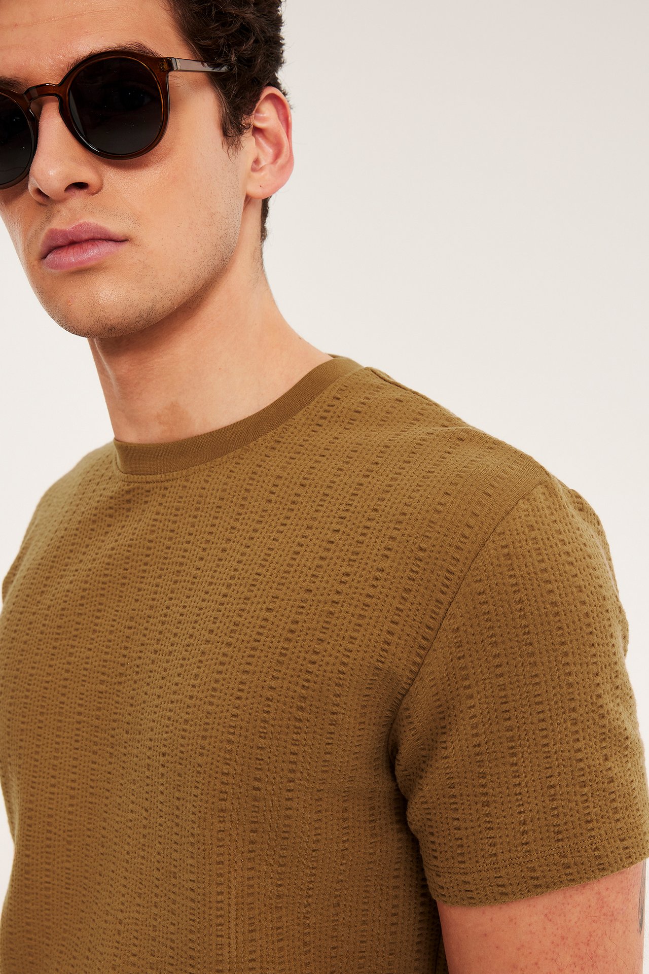 Närbild på en man i en brun strukturstickad O-ringad t-shirt och runda bruna solglasögon.