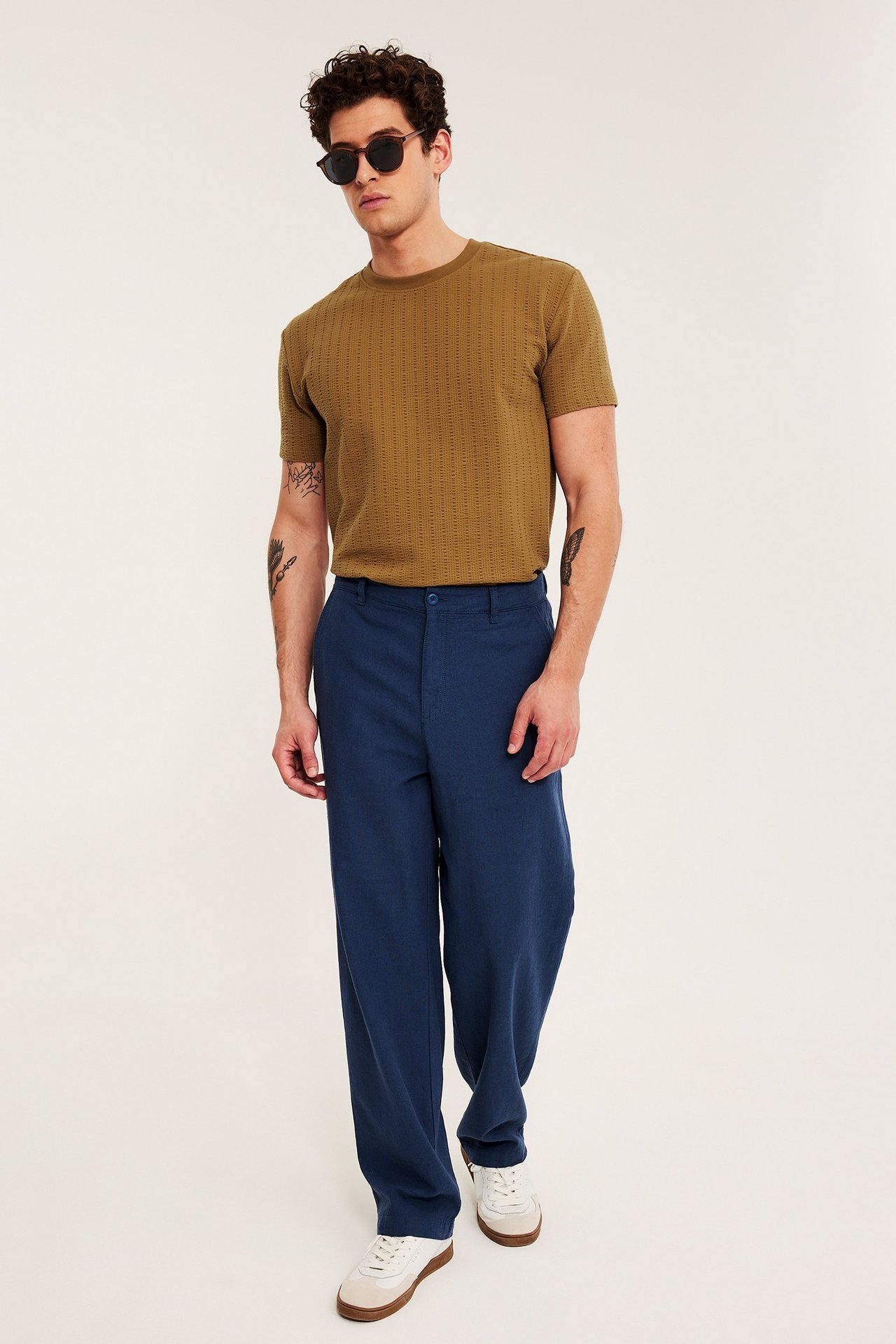 Framifrån: Man i brun strukturerad t-shirt, blåa vida byxor, vita sneakers och solglasögon.