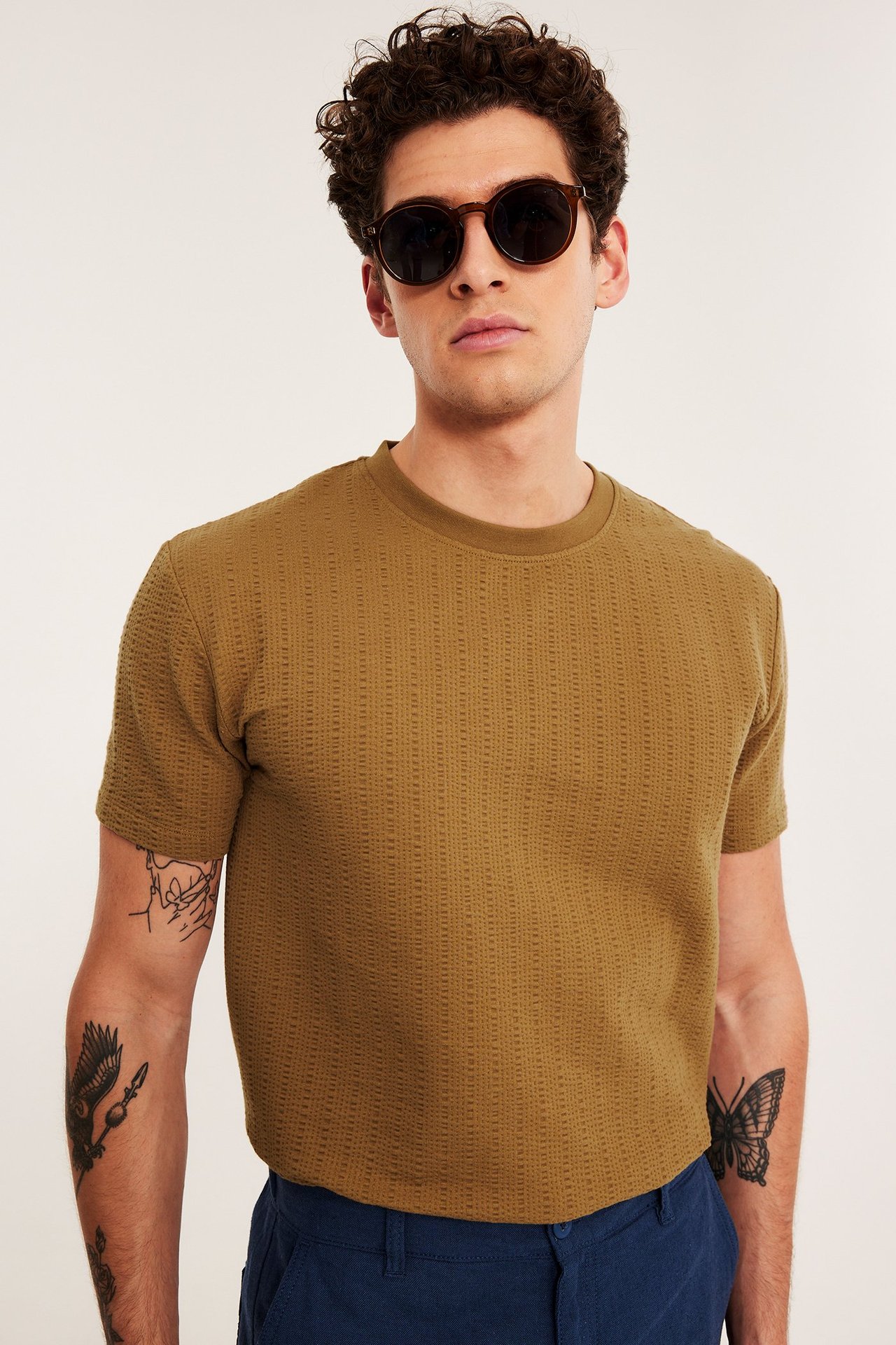 Miehen etukuva ruskeassa, normaalileikkuisessa, O-aukkisessa, struktuurineulotussa t-paidassa, tummissa aurinkolaseissa ja sinisissä housuissa.