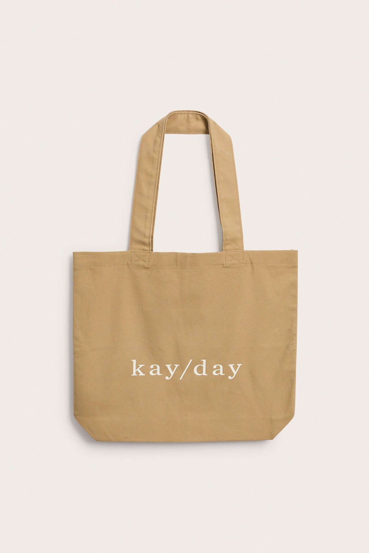 Edestä näkymä beigestä kangaskassista, jossa on valkoinen 'kay/day'-painatus edessä.