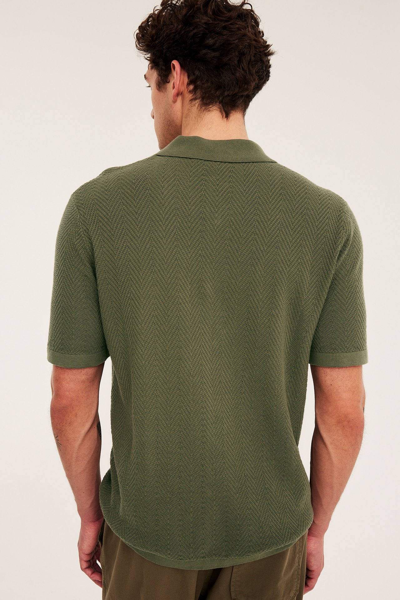 Bakfra, en mann i en grønn strikket piké med diskret fiskebensmønster og grønne bukser.