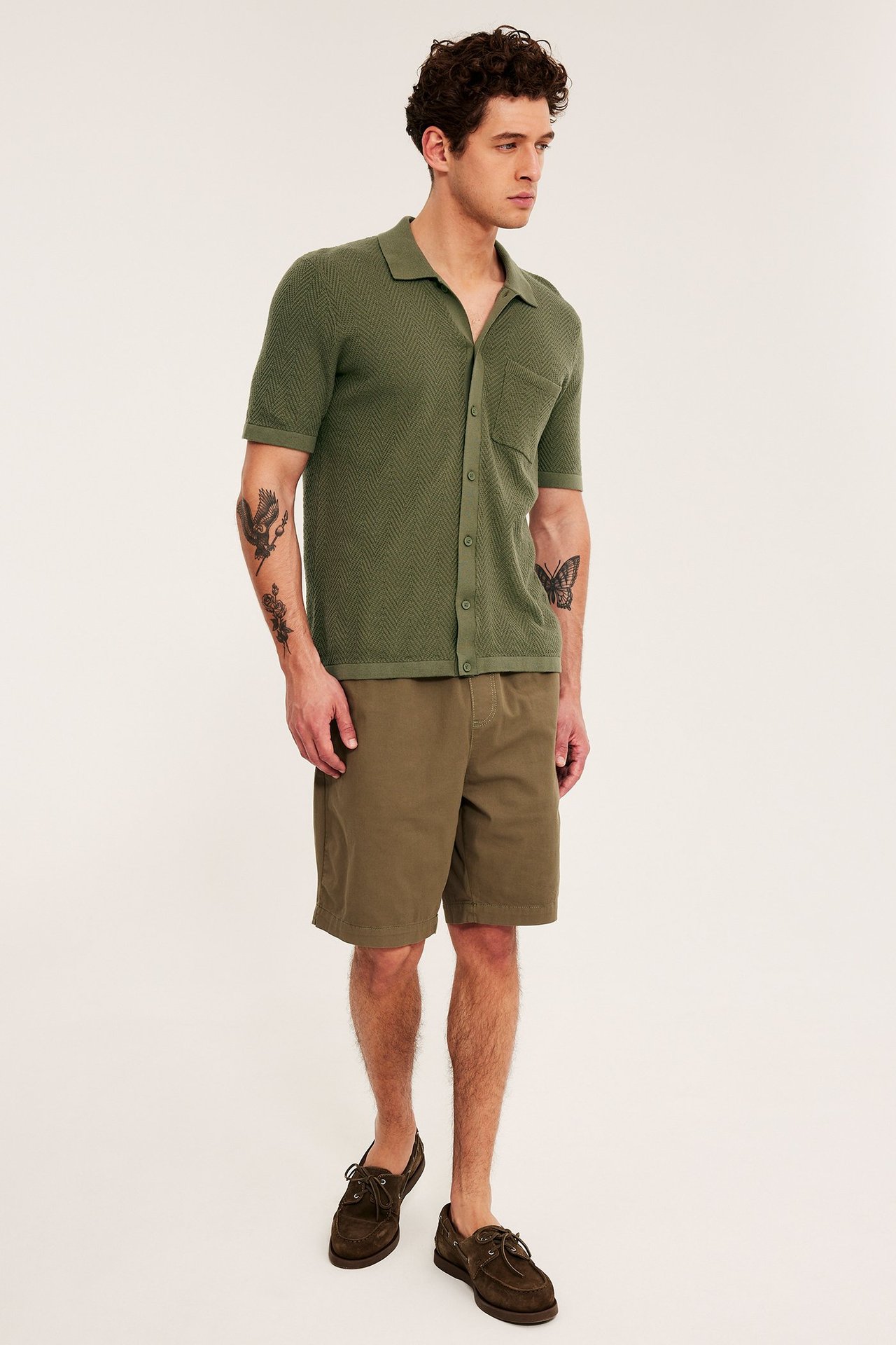 Mann forfra iført en grønn strikket kortermet piké, oliven shorts og brune båtsko.