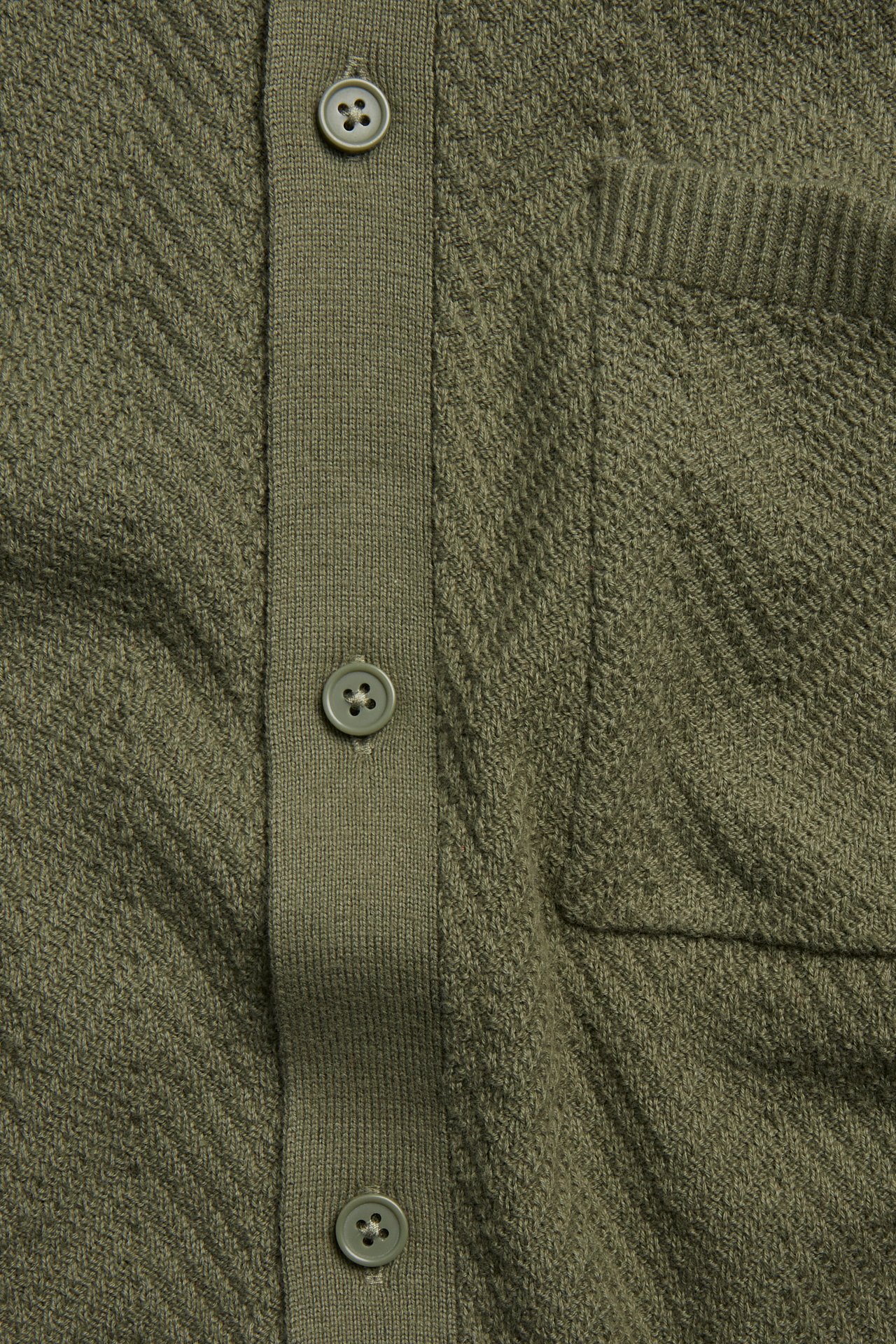 Närbild av en grön stickad pikétröja med chevronmönster, knappslå och bröstficka.