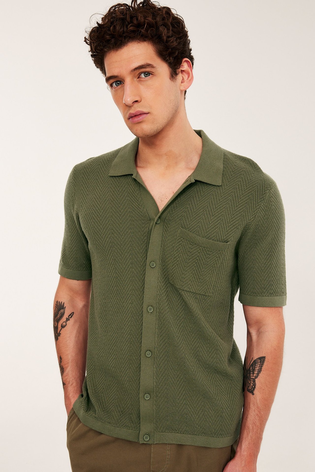 Mężczyzna w zielonej, dzianinowej koszulce polo z krótkim rękawem, we wzór jodełki, z kieszenią i brązowych spodniach.