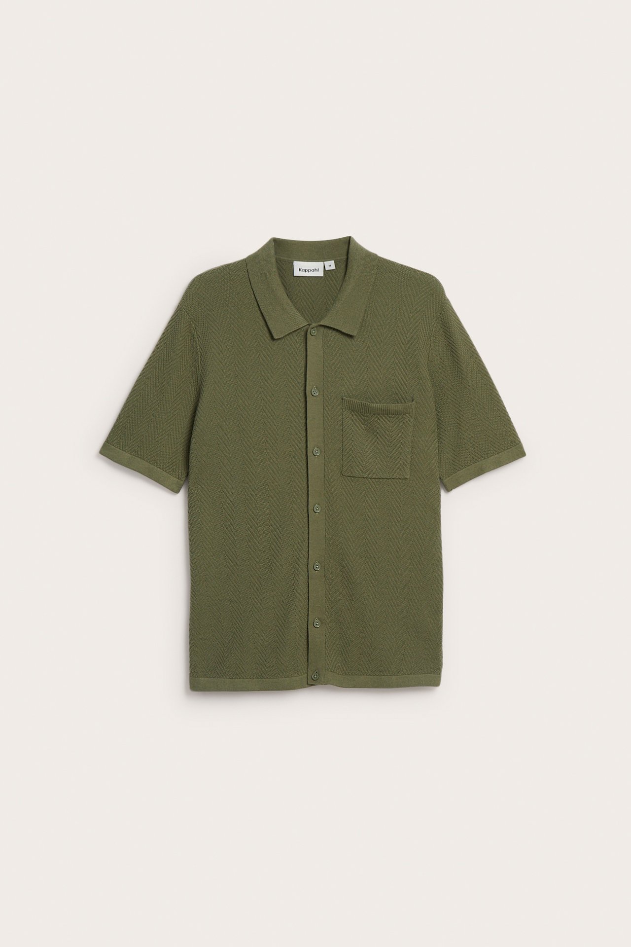 Framsida av en grön stickad kortärmad pikétröja med diskret chevronmönster, helknäppning och bröstficka.