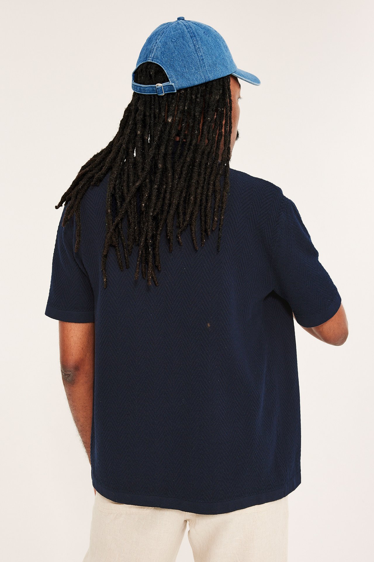 Mann bakfra i en mørkeblå strikket kortermet pikétrøye, blå denimcaps og lyse beige bukser.