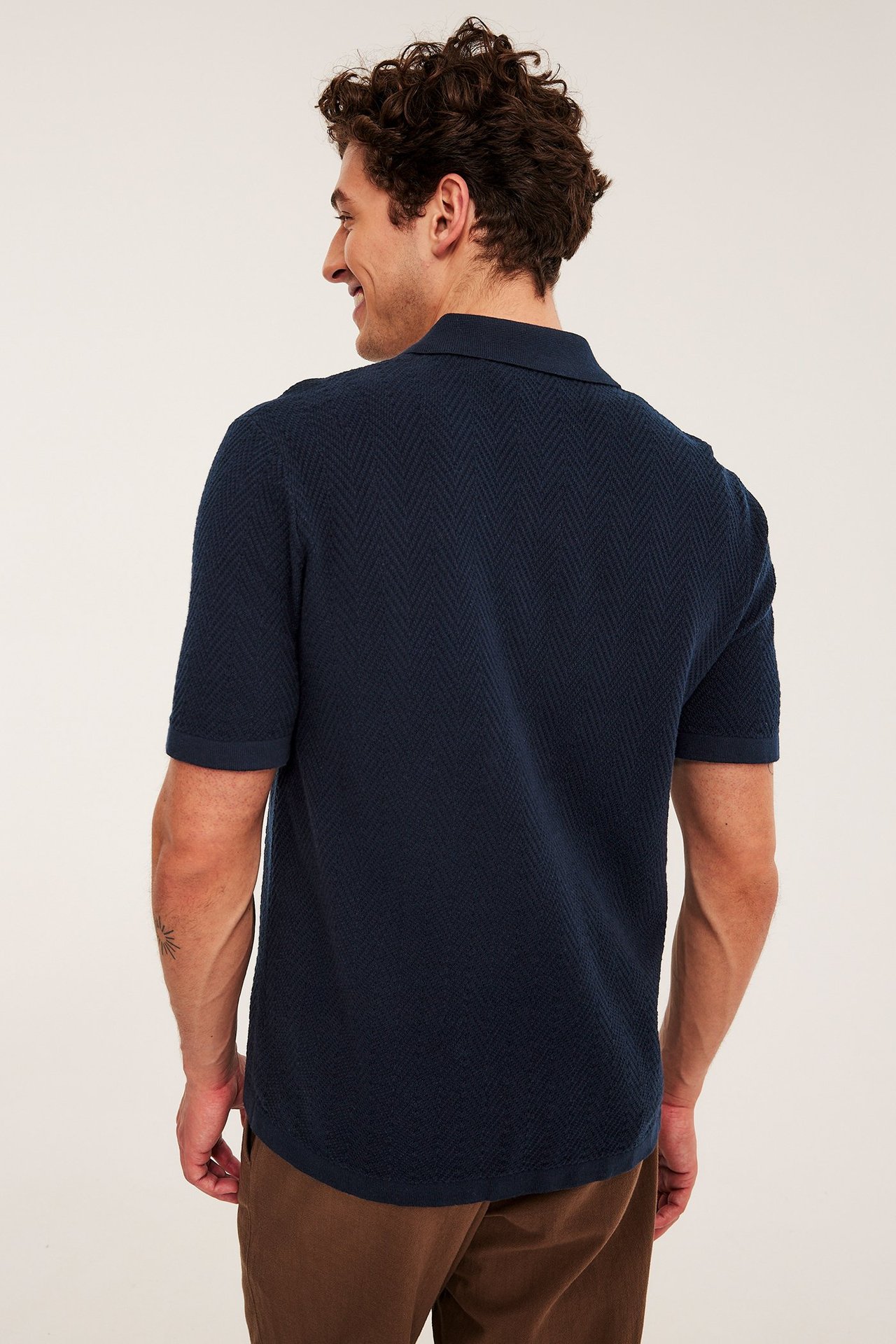 Mann bakfra, iført en mørk blå strikket piké med teksturert mønster og brune bukser.