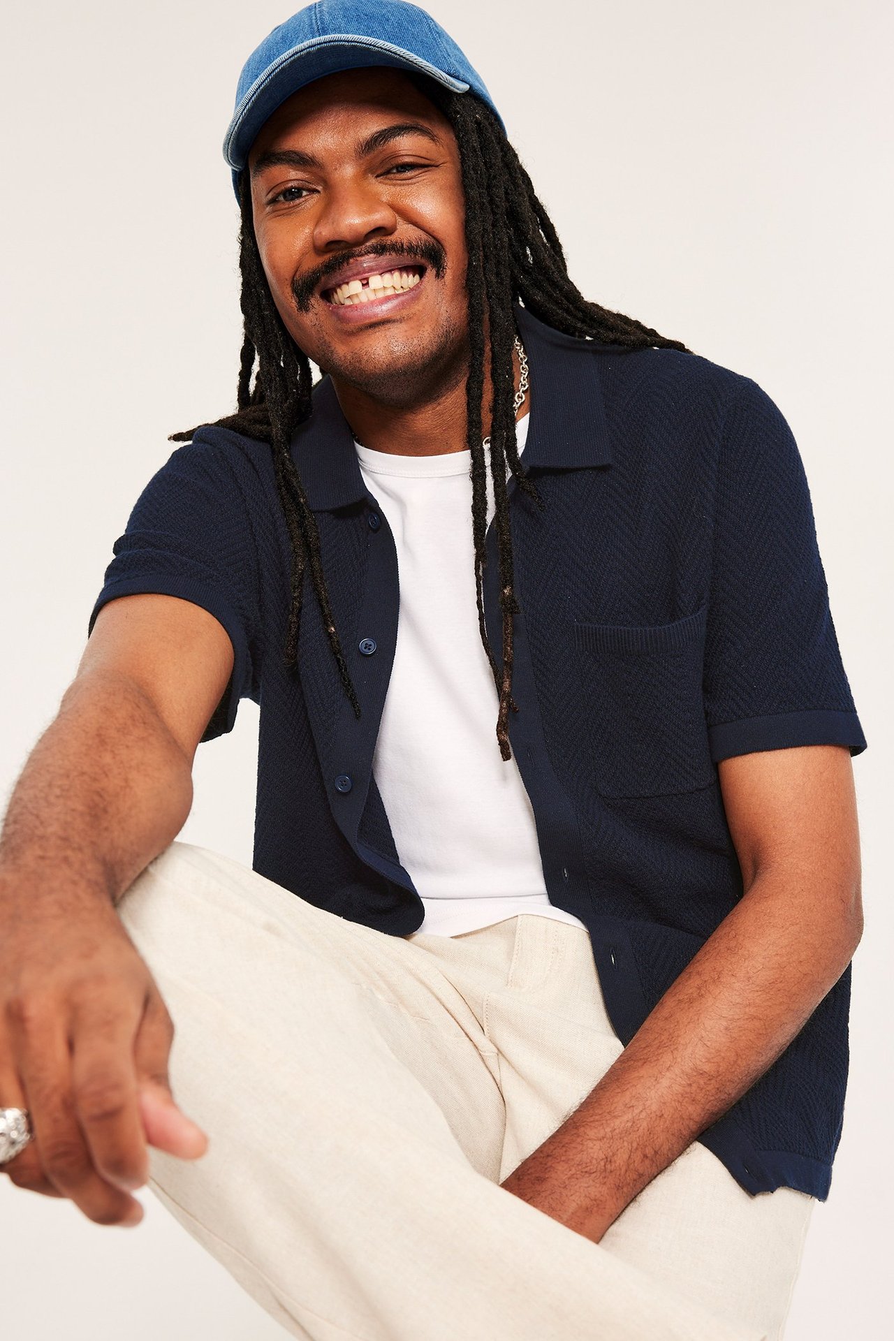 Frontvy: Mann har på seg mørkblå strikket kortermet poloskjorte, hvit t-skjorte, beige bukser, blå dongerikeps.