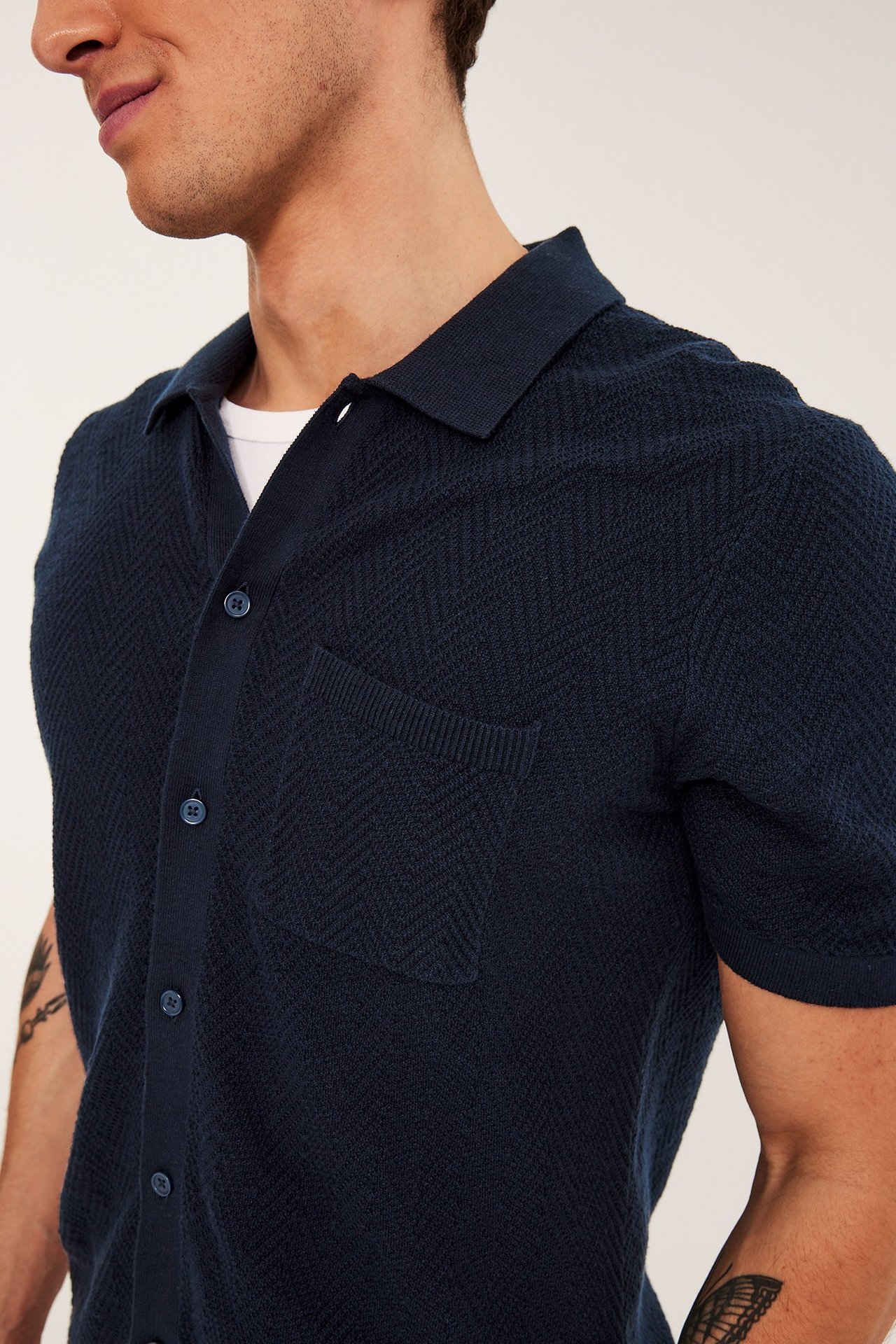 Nærbilde av en mann i en mørkeblå kortermet strikket poloskjorte med brystlomme over en hvit t-skjorte.