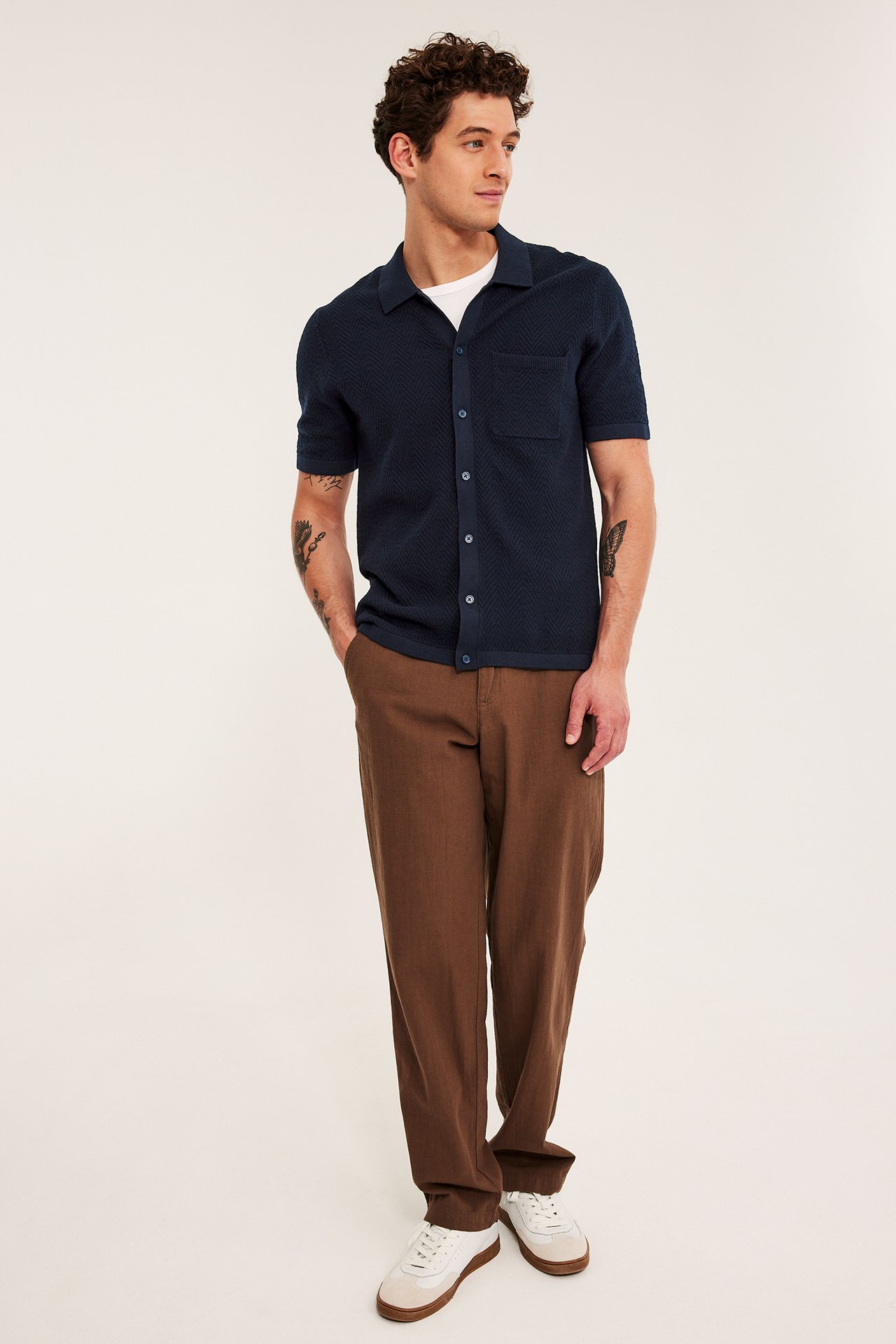 Mann, forfra, i mørkeblå strikket kortermet polotrøye, brune bukser og hvite joggesko.