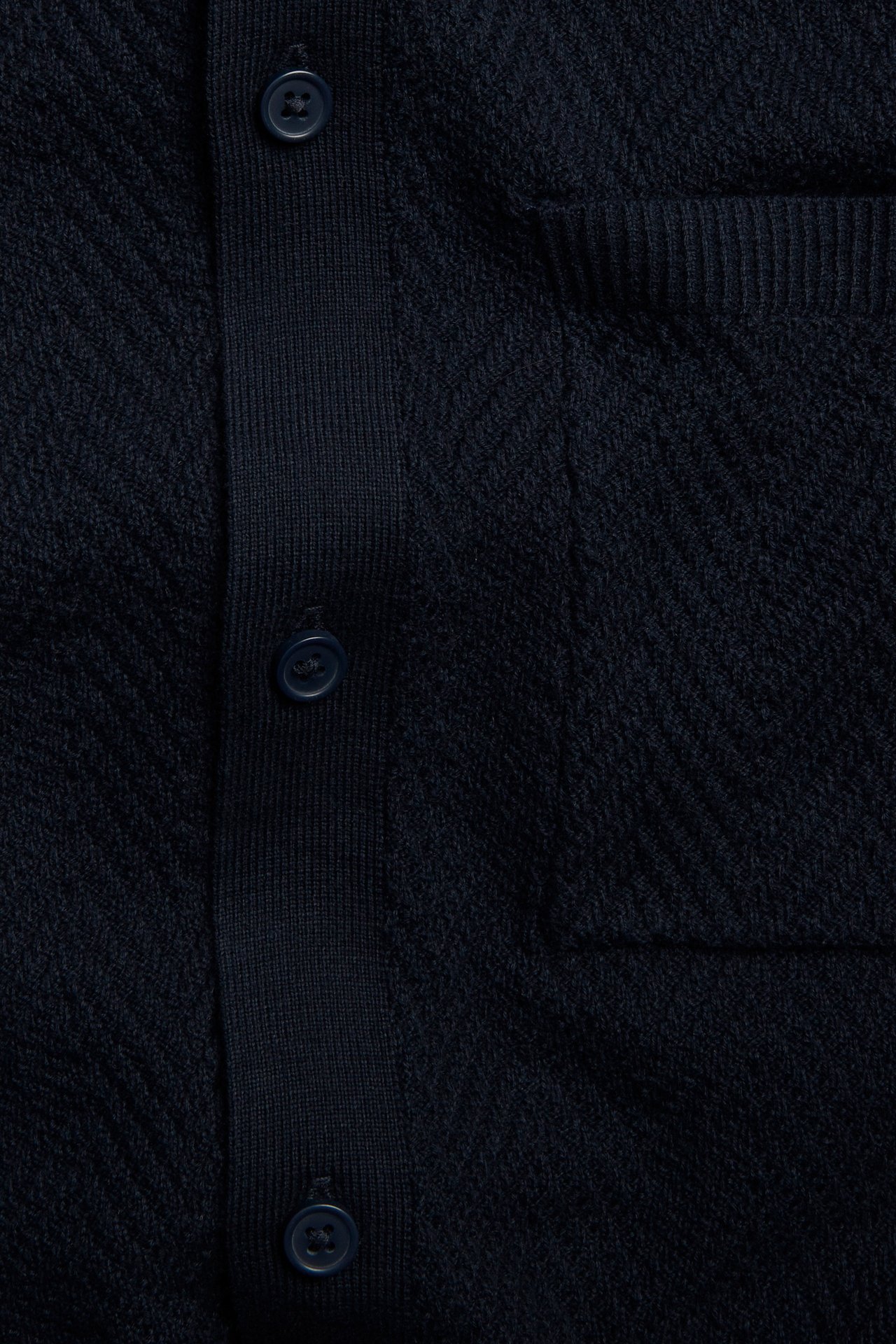 Närbild framifrån på en mörkblå stickad pikétröja med knappslå och bröstficka.