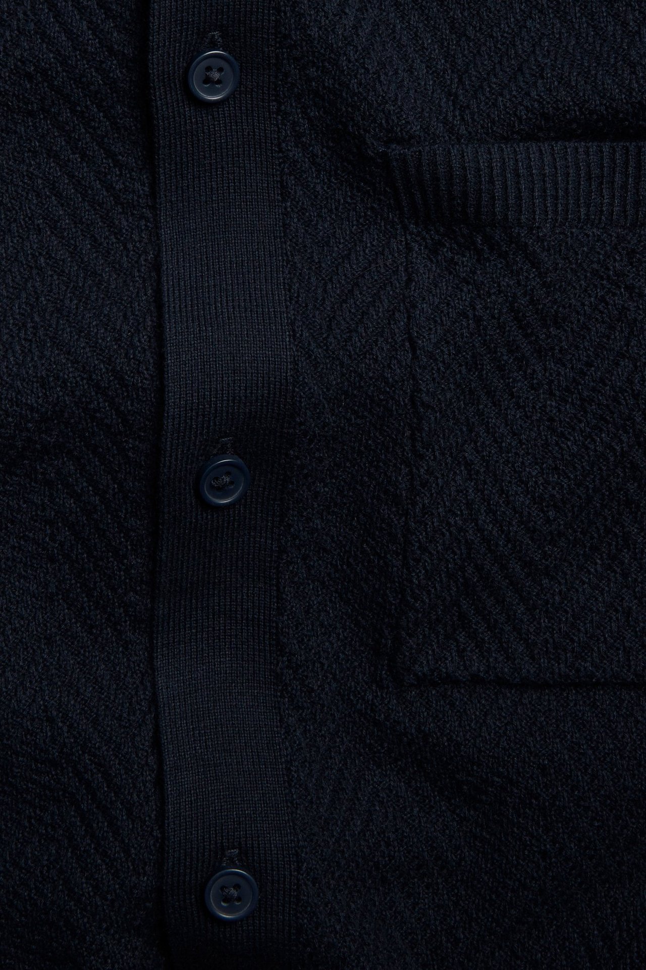 Närbild framifrån på en mörkblå stickad pikétröja med knappslå och bröstficka.