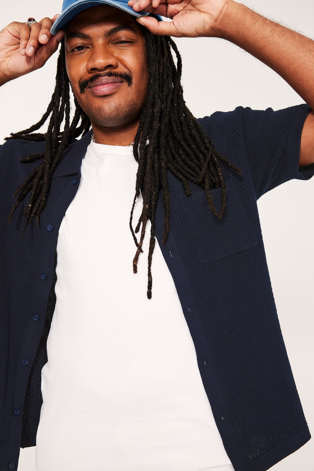 Forfra: mann i mørkeblå kortermet strikket skjorte med brystlomme over hvit t-skjorte, holder blå caps.