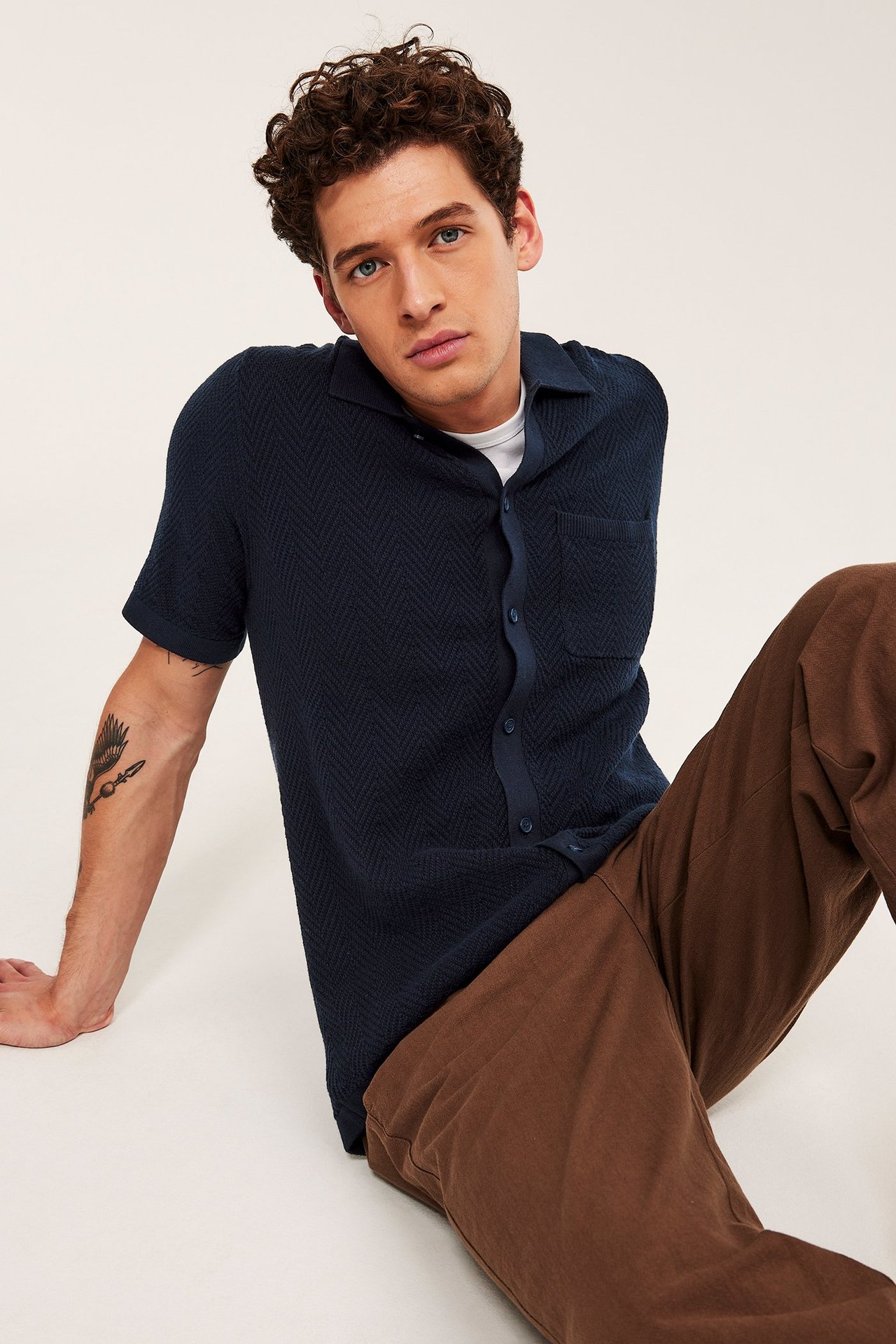 Mężczyzna nosi granatową dzianinową koszulkę polo z krótkim rękawem, kieszonką, guzikami i brązowe spodnie.