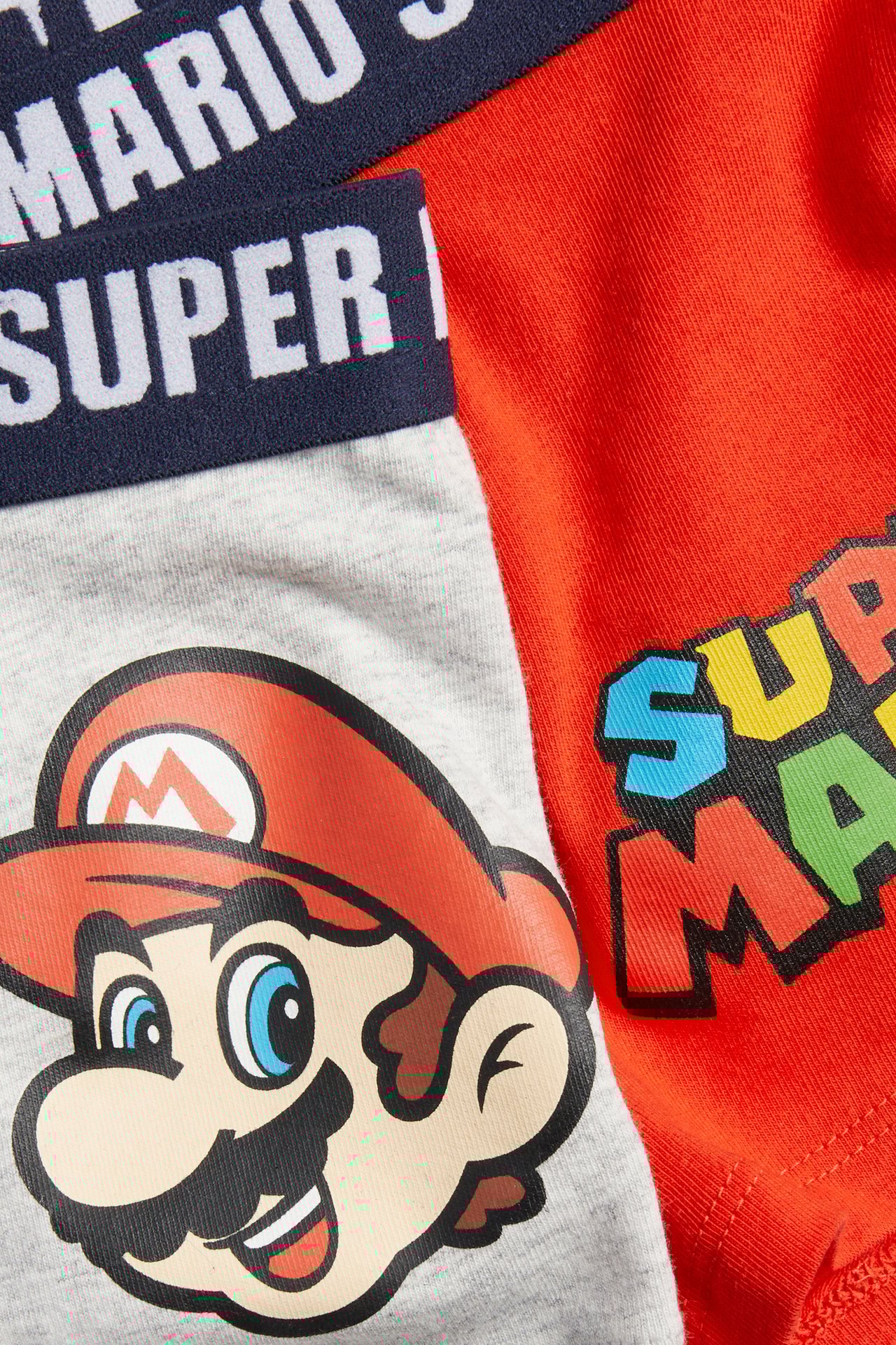 Närbild: två par barnkalsonger i bomull. Ett gråmelerat par med Super Mario-ansikte, ett rött par med Super Mario-logotyp.