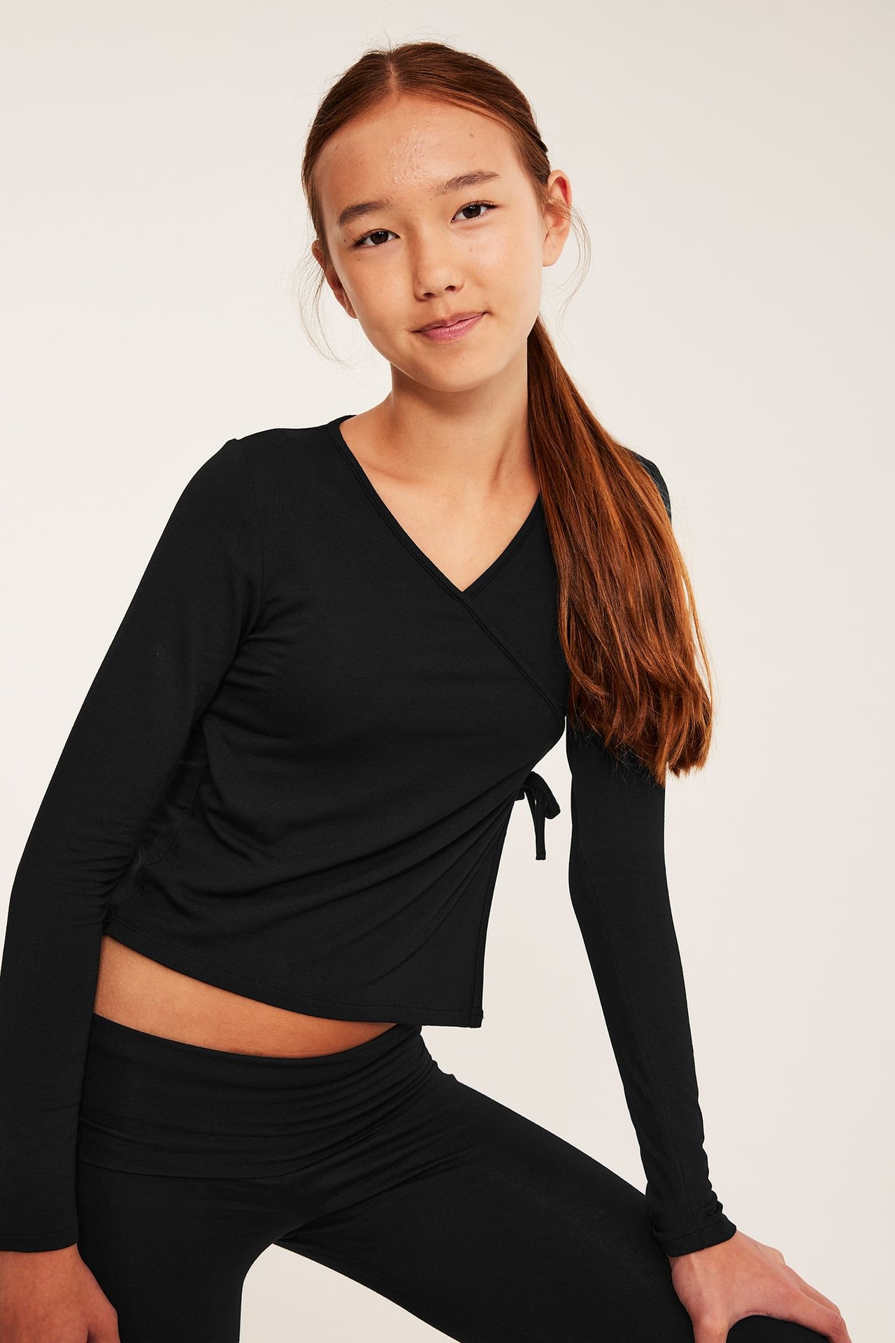 Frontvy av barn i svart långärmad omlott-topp med knytband och svarta rynkade leggings.