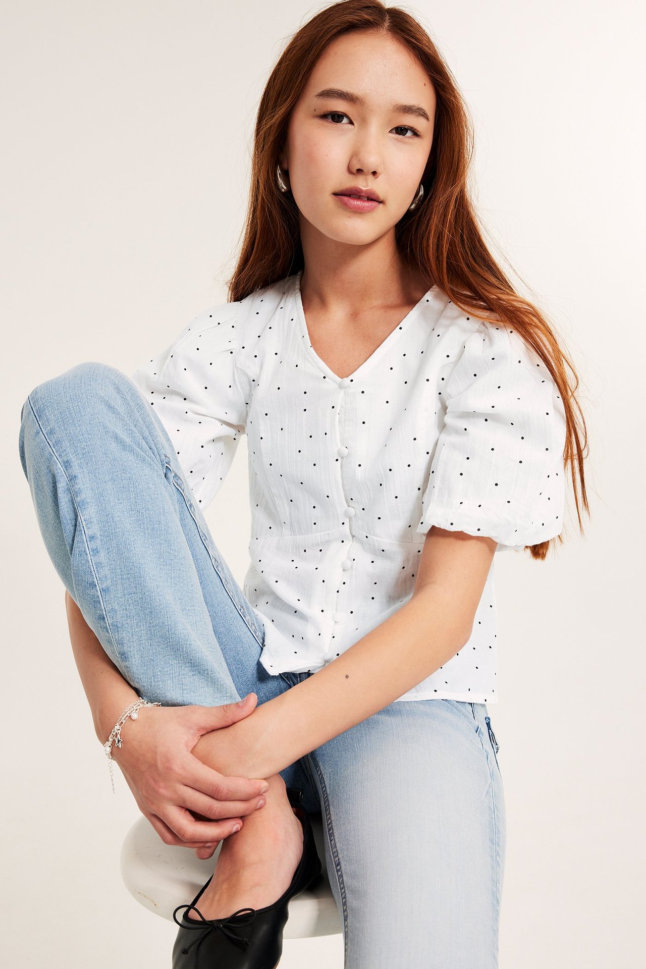 Widok z przodu dziecka w białej bluzce w kropki z bufiastymi rękawami, jasnoniebieskich jeansach i czarnych balerinach.