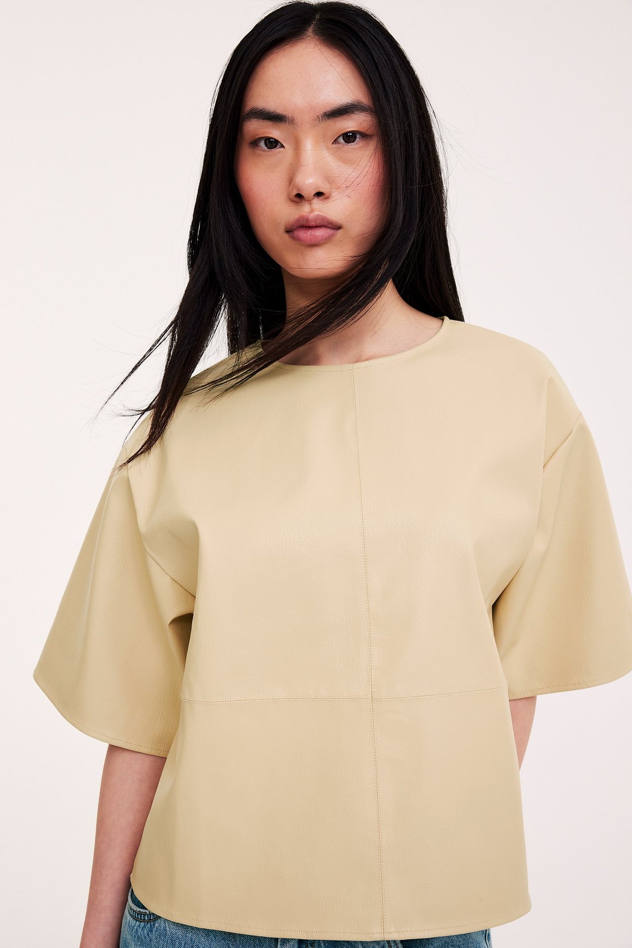 Edestä: Nainen yllään väljä vaaleankeltainen lyhythihainen tekonahkatoppi ja näkyvät siniset farkut.