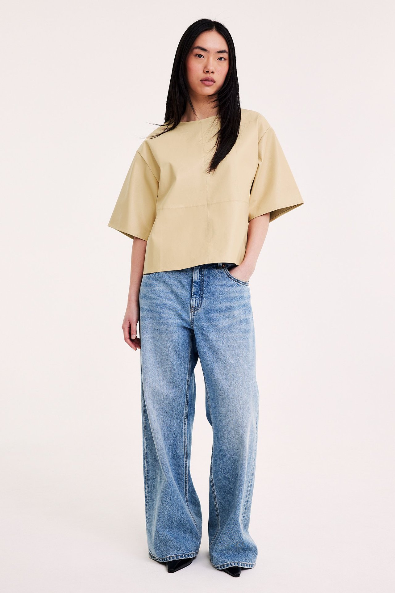 Edestä: nainen ylisuuressa vaaleankeltaisessa keinonahkatopissa, vaaleansinisissä leveissä farkuissa, mustissa kengissä.