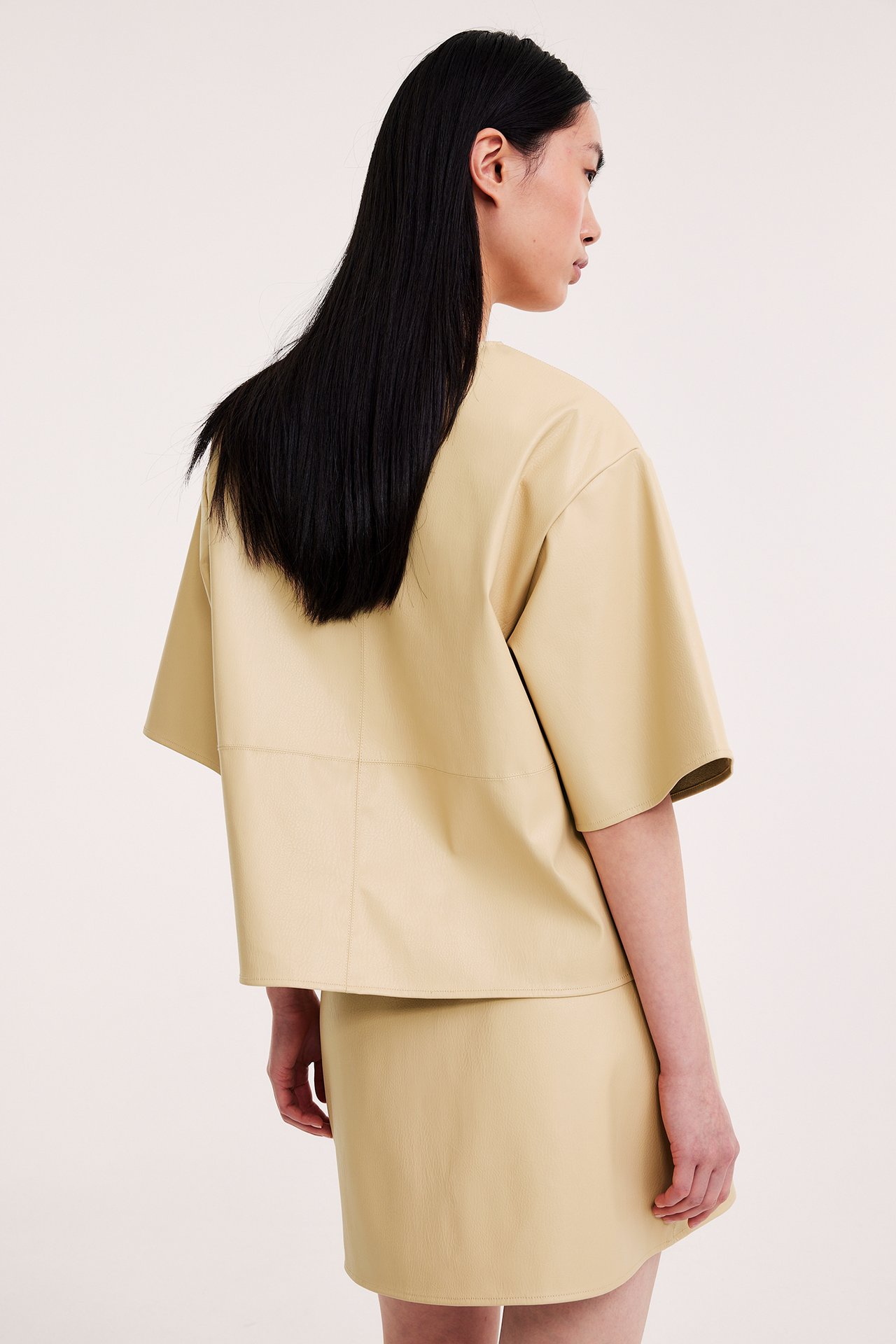 Sivu-takanäkymä: nainen yllään vaaleankeltainen lyhyt keinonahkahame ja vastaava väljä lyhythihainen toppi.